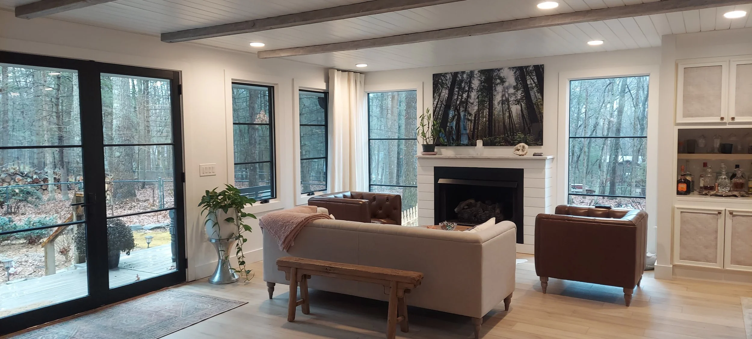 Living room with large windows, a fireplace, a beige sofa, brown leather armchairs, a potted plant, a wooden bench, a gallery wall with a forest scene, and a built-in cabinet with liquor bottles, overlooking a wooded backyard.