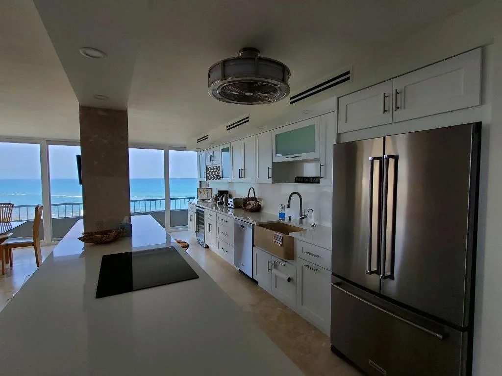 Modern kitchen with white cabinets, stainless steel refrigerator, and view of ocean through large windows.