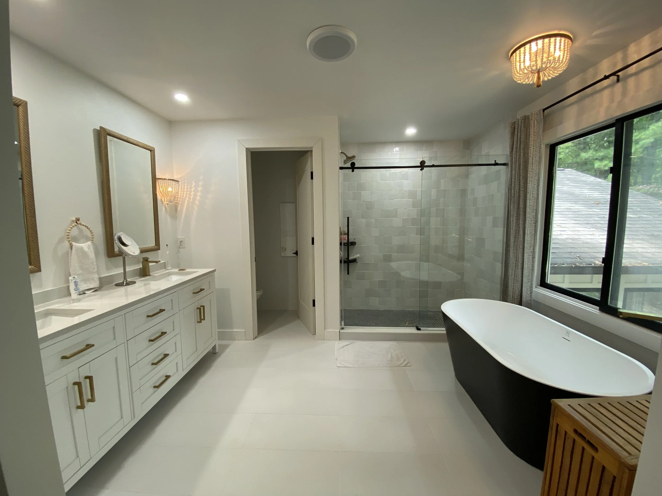 Modern bathroom featuring a white vanity with gold handles, a large mirror, a freestanding bathtub, a walk-in tiled shower with a sliding glass door, and a large window with curtains showing greenery outside.