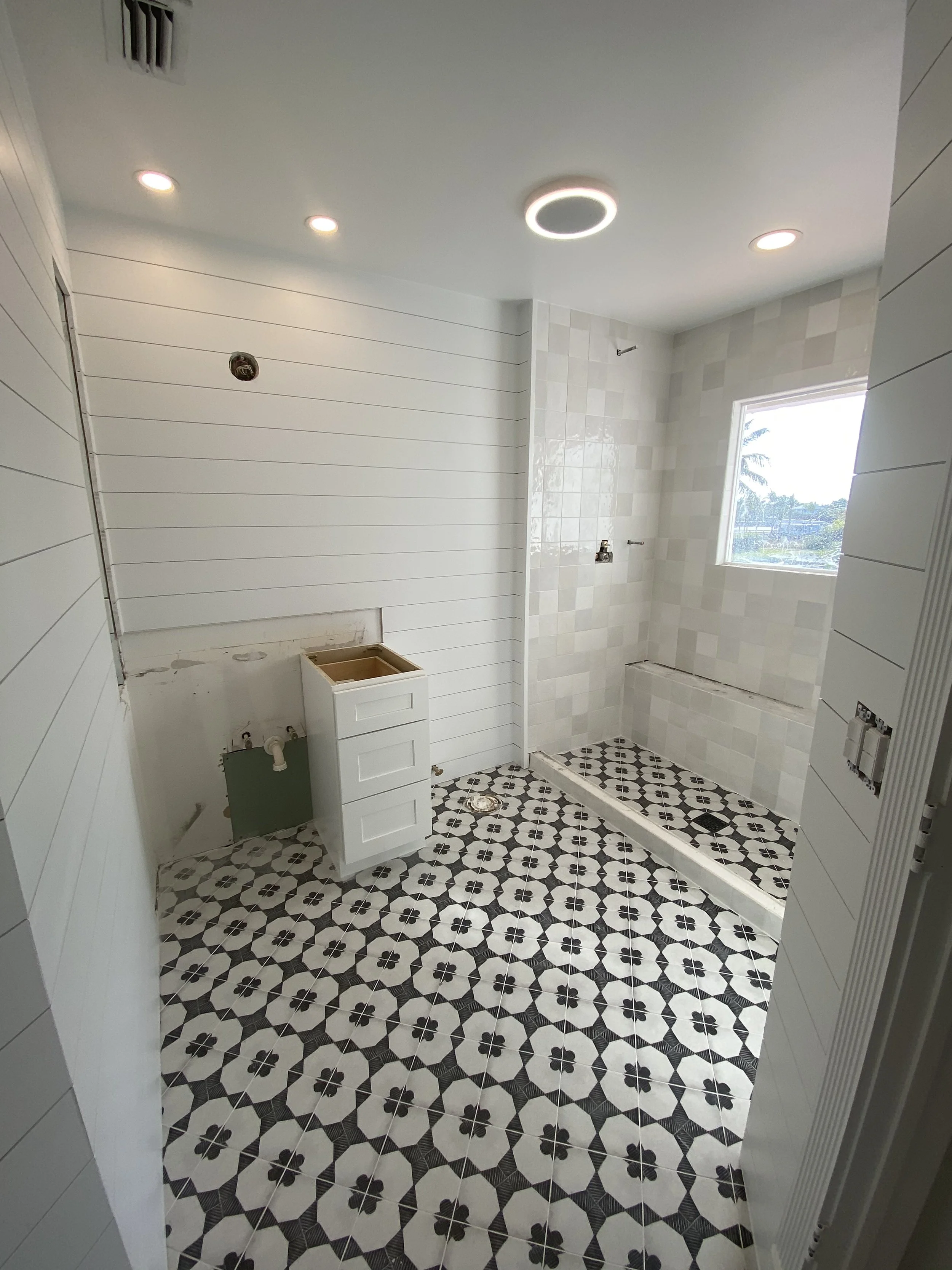 A bathroom under construction with patterned black and white floor tiles, white shiplap walls, a window, and partial plumbing fixtures, including a vanity base and shower area.