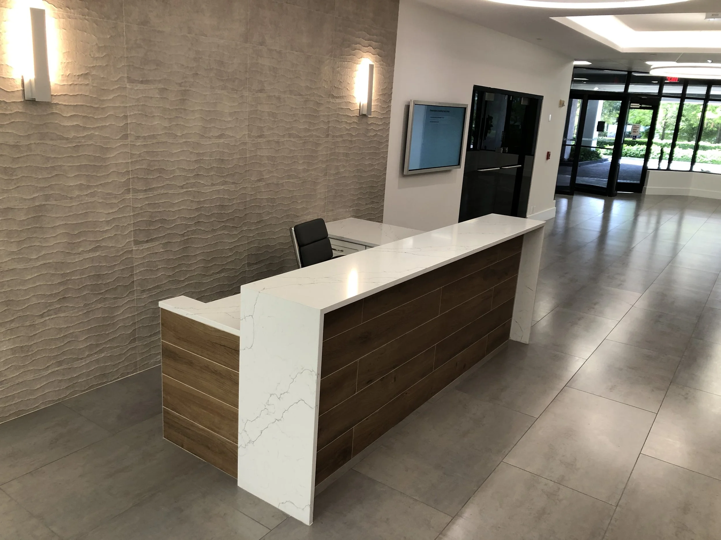 Empty modern reception desk with marble top and wooden front panel in a brightly lit lobby area, with a textured beige wall and a wall-mounted television.