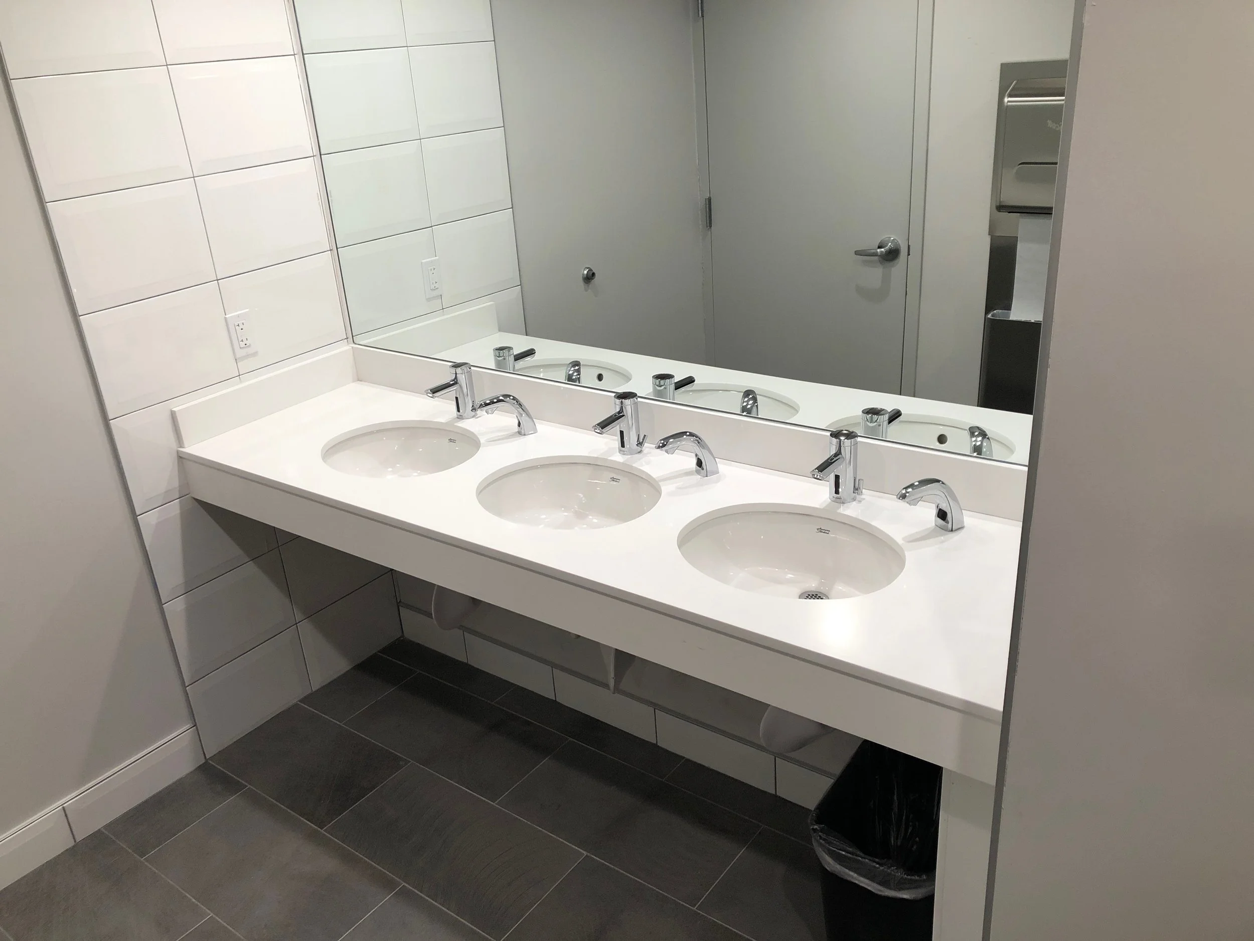 A restroom sink area with three white sinks, a large mirror above, showing a bathroom door, a paper towel dispenser, and trash can.
