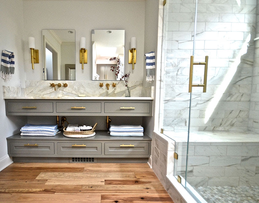 Modern bathroom with a gray vanity, three mirrors, and gold fixtures; towels and a small decorative branch on the counter; walk-in shower with glass enclosure and marble tiles; hardwood floor.