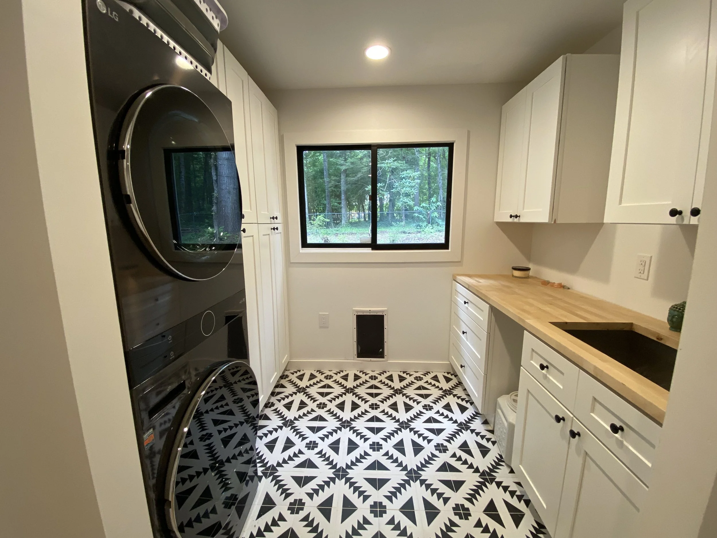 A laundry room with black and white patterned tile flooring, white cabinets, a wooden countertop, a black undermount sink, a window showing a forest outside, a stacked washer and dryer, and a vent or heater on the wall.
