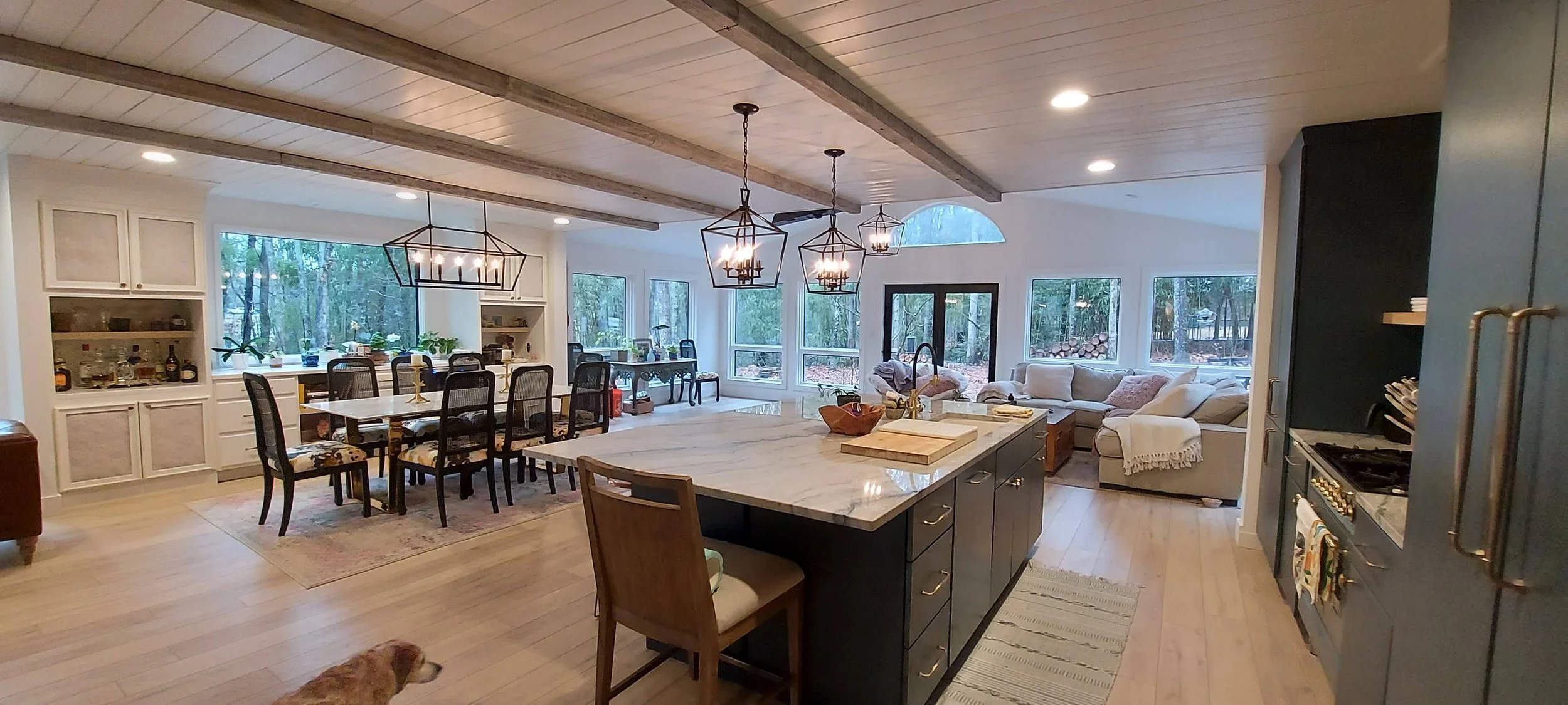 Open-concept kitchen and living area with large windows, white cabinetry, black dining table with chairs, island with marble countertop, and beige sectional sofa, with a view of a wooded backyard.