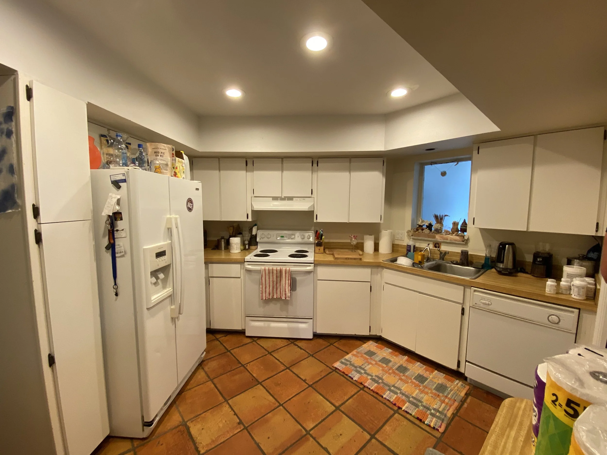 A kitchen with white cabinets, a white stove, and a white refrigerator. The countertop has various kitchen items, and there is a small window above the sink. The floor is tiled with reddish-brown tiles, and a colorful rug is in front of the sink.