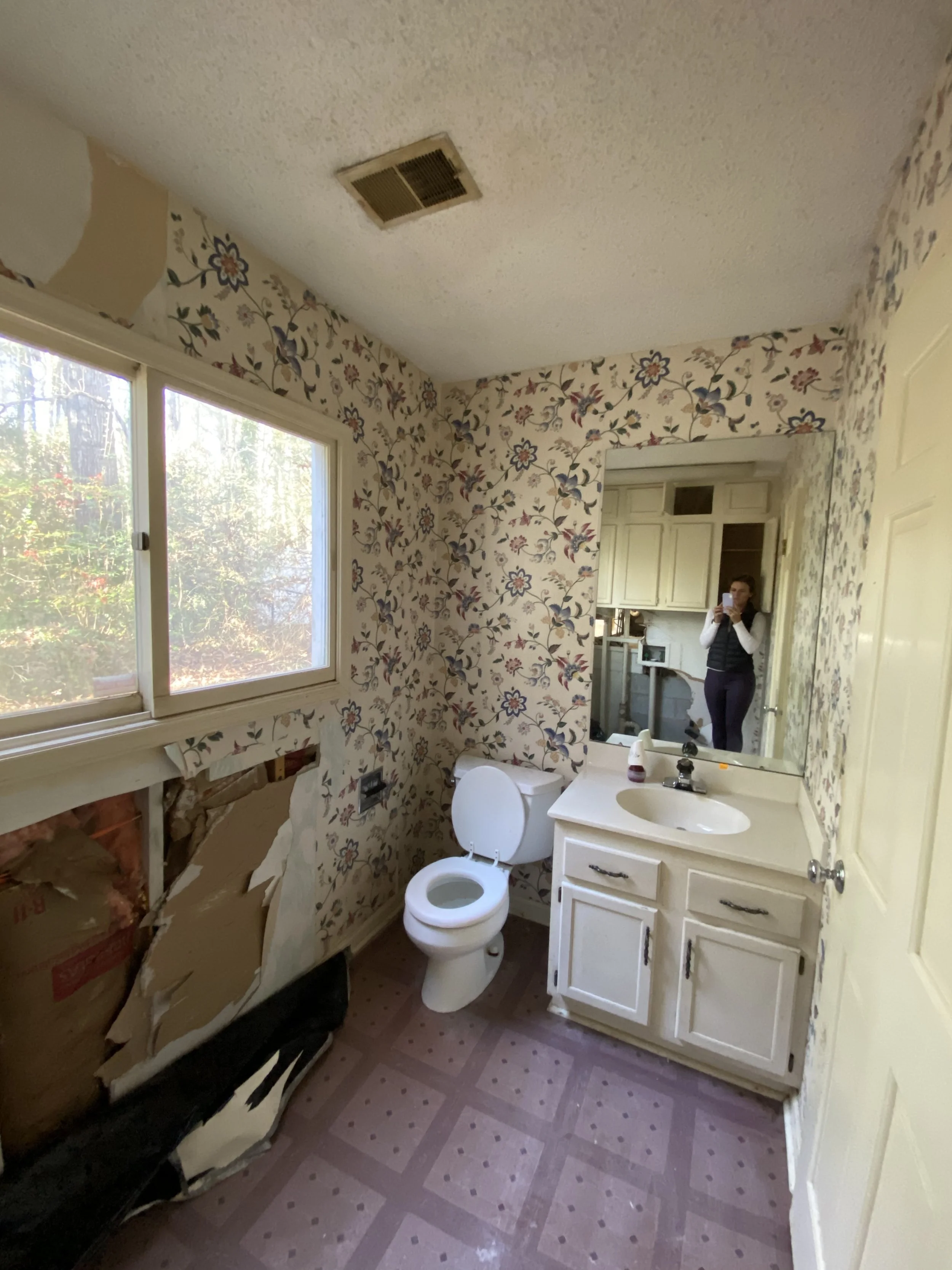 A bathroom with floral wallpaper, a window, a toilet, a sink with a mirror, and a woman taking a selfie in the mirror. The wall beneath the window is damaged with exposed insulation.