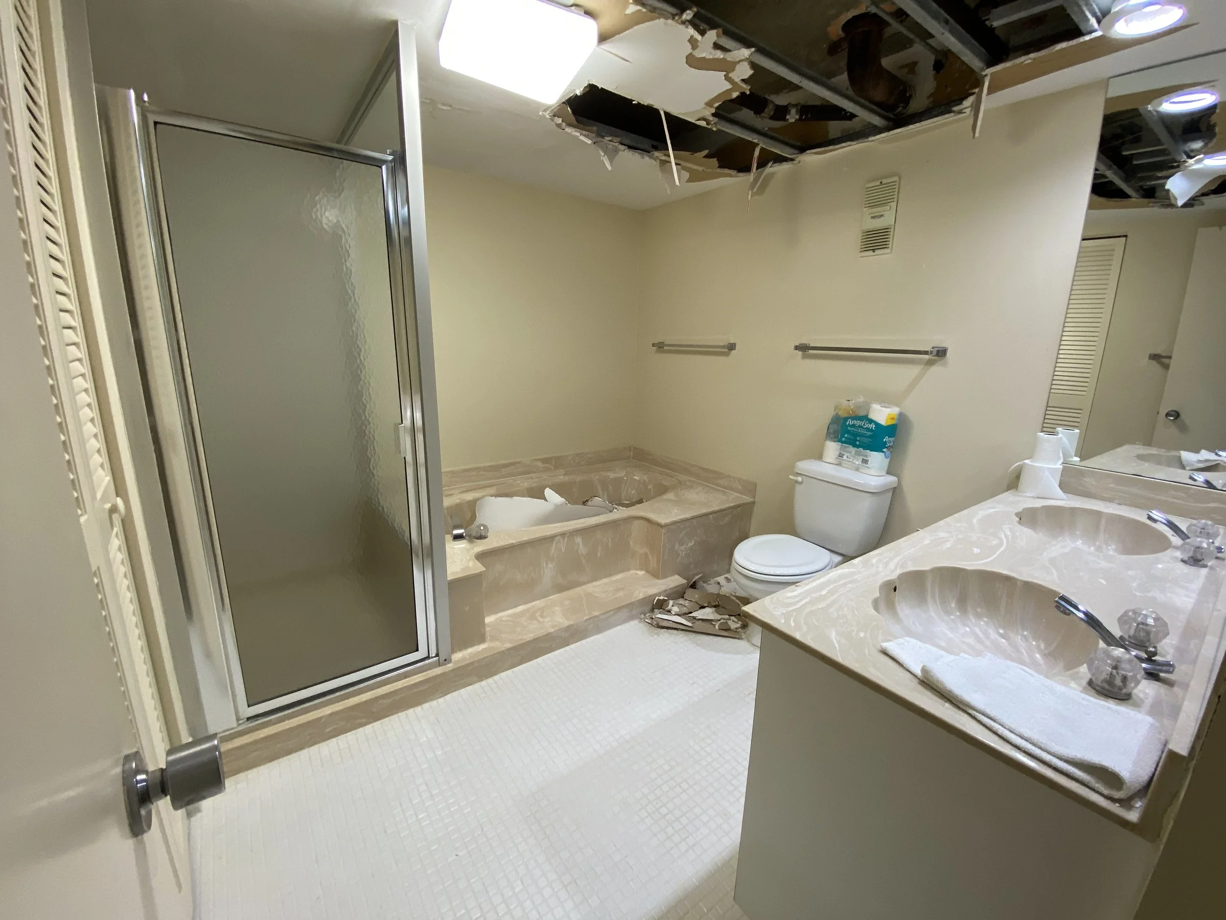 Bathroom under renovation with ceiling damage, missing tiles on the floor, and construction debris. There is a shower stall, a bathtub, a toilet, and a double sink vanity with a mirror. The ceiling has exposed pipes and drywall, some of which is hang