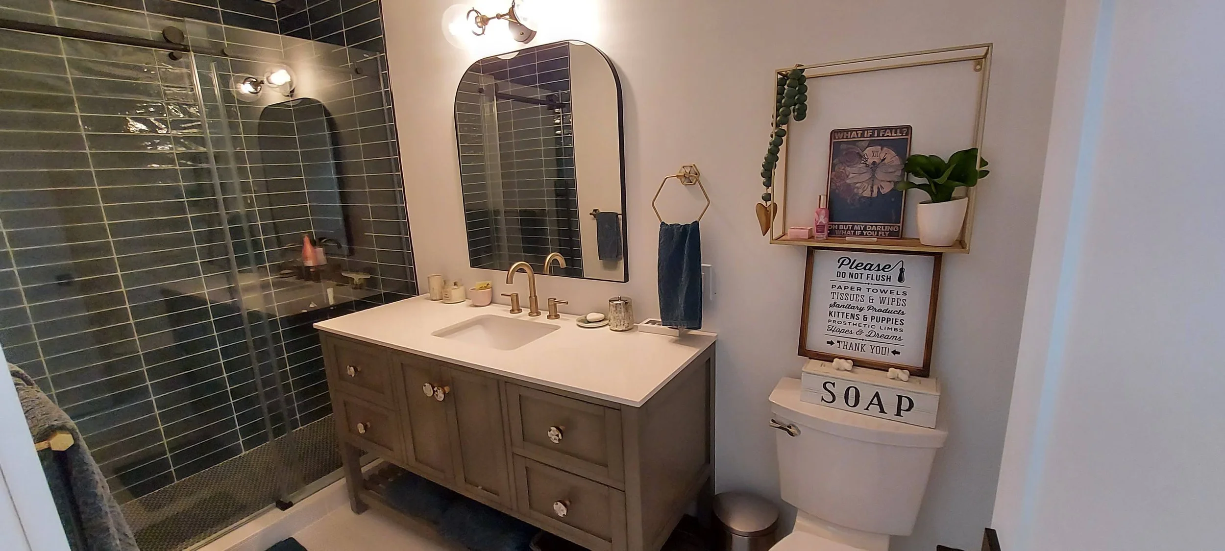 A modern bathroom with a gray vanity, white countertop, and gold fixtures. There is a large mirror above the vanity, a glass-enclosed shower with black tiles, and decorative items including plant and framed art on the wall, along with a decorated toi