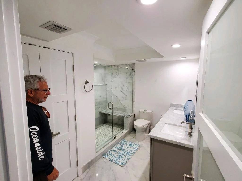 A man standing in a modern bathroom with marble floors, a glass-enclosed shower, white walls, and a double sink vanity with toiletries and a large mirror.