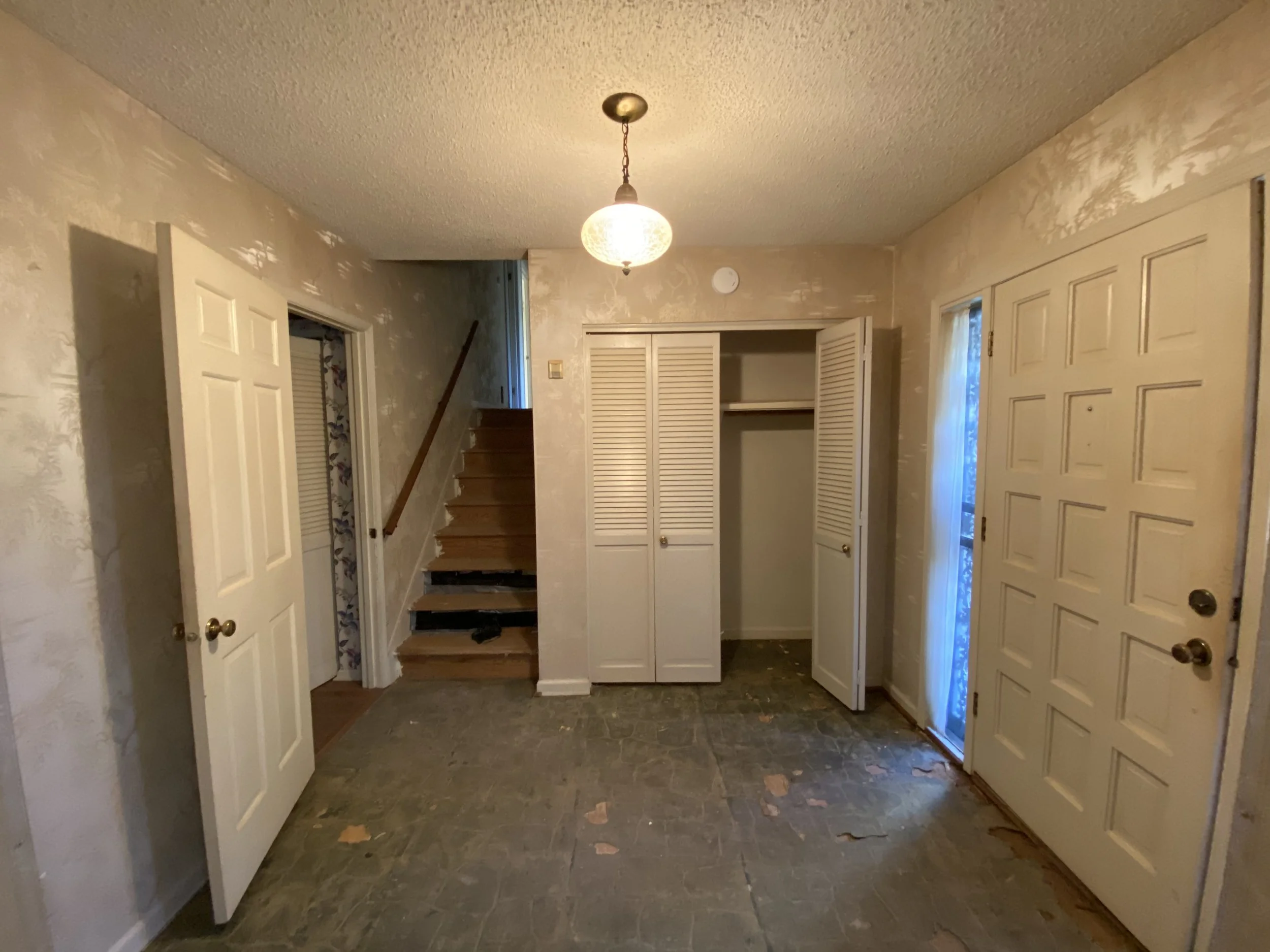 Empty entryway with open closet doors, staircase, and a door with a curtain, unfinished walls, ceiling light, and a stained floor.