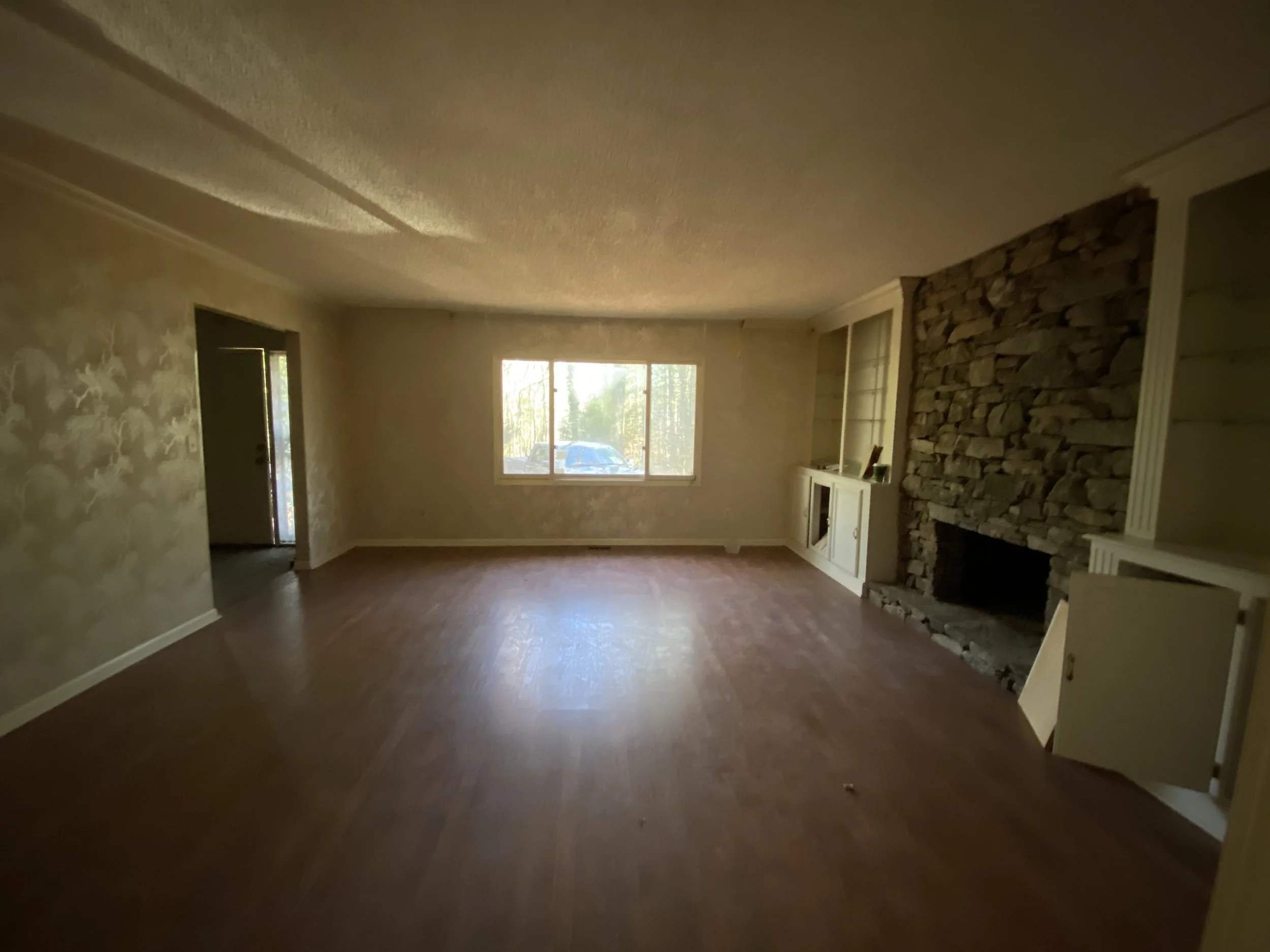 Empty living room with wood flooring, a stone fireplace, built-in cabinets, and a large window with trees outside.