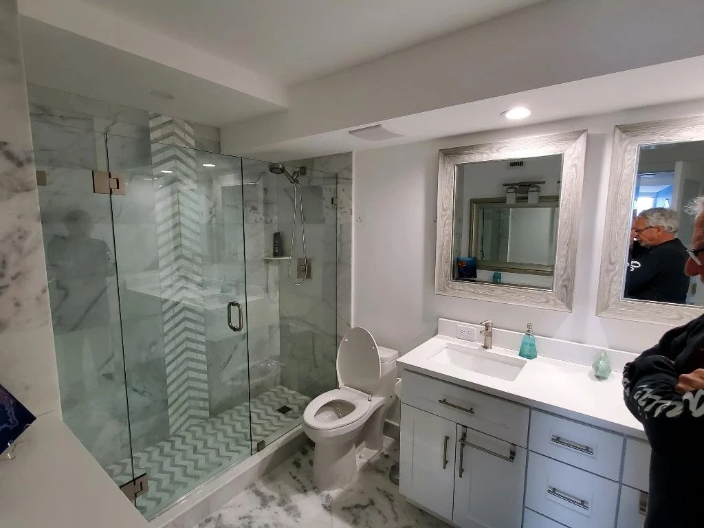 Modern bathroom with glass-enclosed shower, white vanity with sink, and large mirrors. Two people are visible in the reflection of the mirrors.