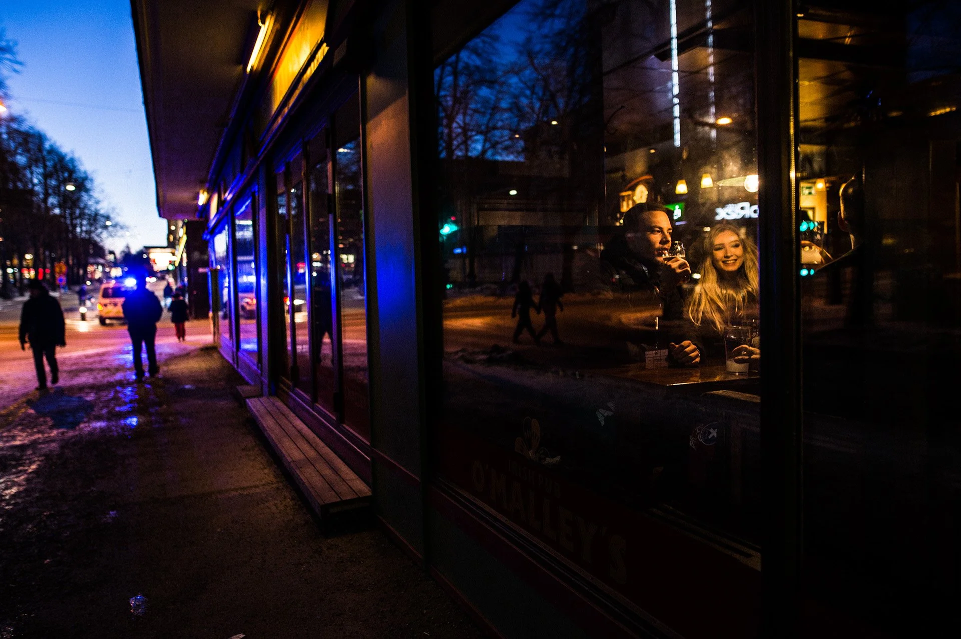 Yksityiskohtainen kuva iltavalaistuneesta kahvilasta, jonka ikkunasta näkyy kaksi ihmistä nauttimassa juomia, taustalla ulkona kävelee ihmisiä ja liikennevaloja.