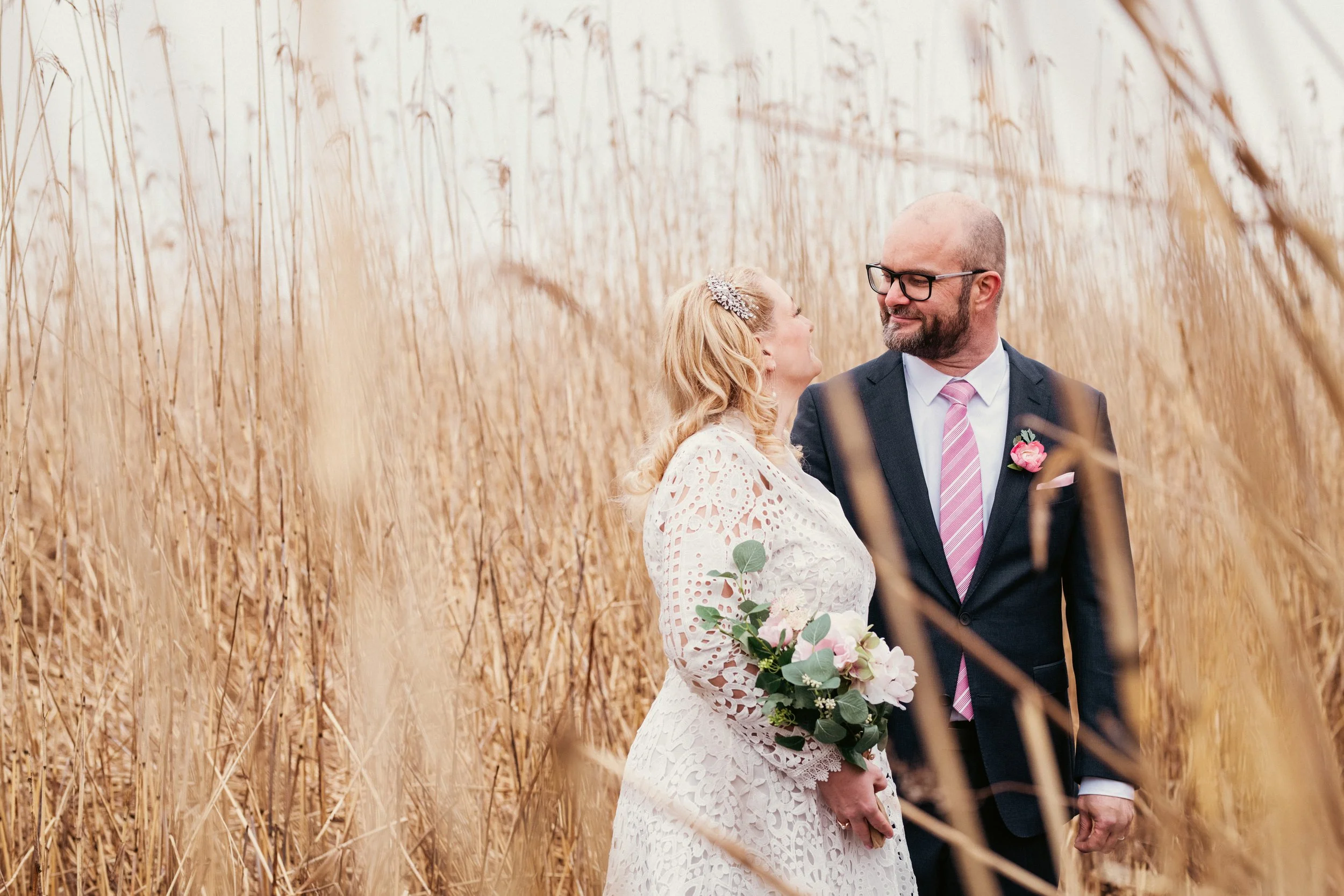 Hääpari seisoo heinän seassa, molemmat hymyilevät ja katsovat toisiaan, nainen pitää kukkakimppua ja mies on puketunut pukuun.