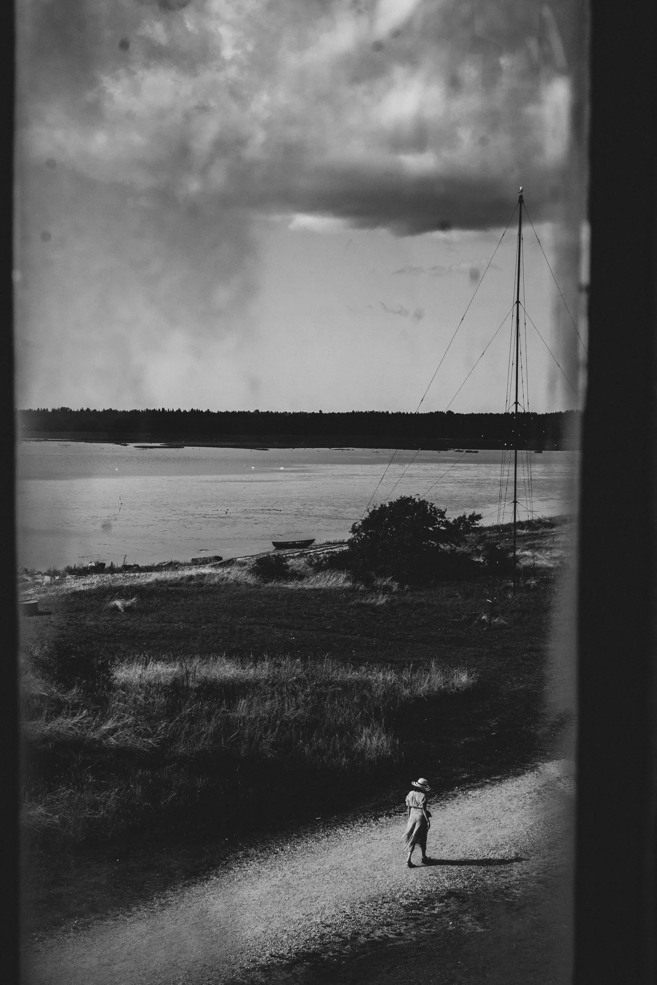 Näkymä rannasta, jossa on vene ja purjehdussatama, näkyy taustalla. Edessä kävelee nainen hatun kanssa polulla.