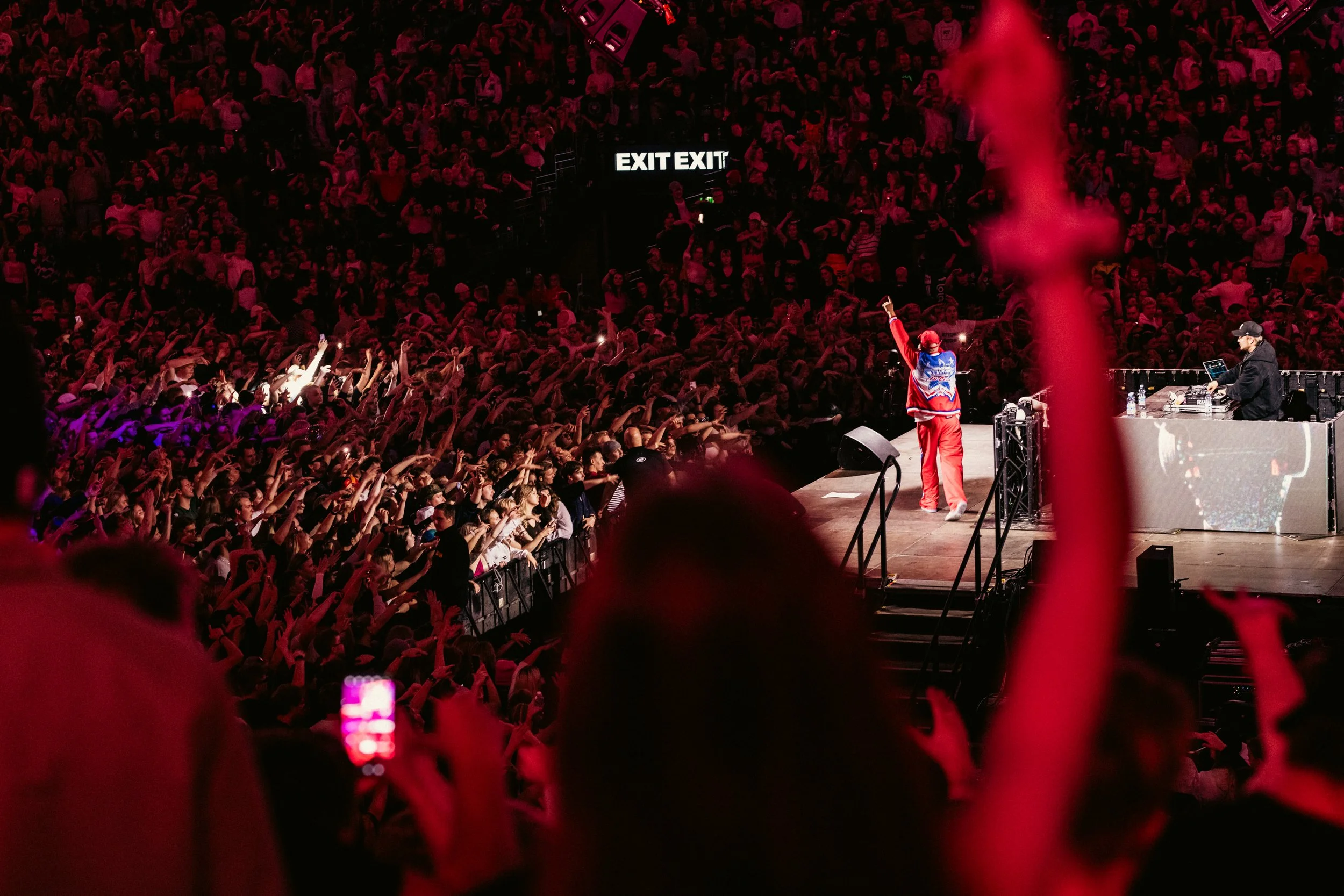 Näytöskonsertti täpötäydessä areenassa, artisti esiintyy lavalla, taustalla kuului exit-merkki, yleisö heiluttaa käsiä ja tallentaa tapahtumaa kännyköillä.