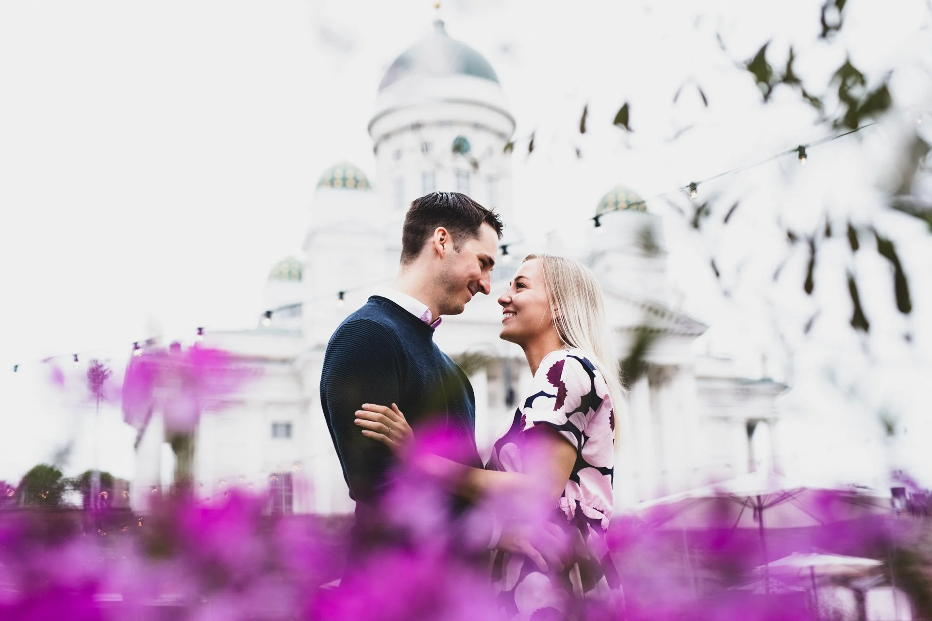 Pari hymyilevät ja katsovat toisiaan läheltä, taustalla valkoinen linna ja taivasta