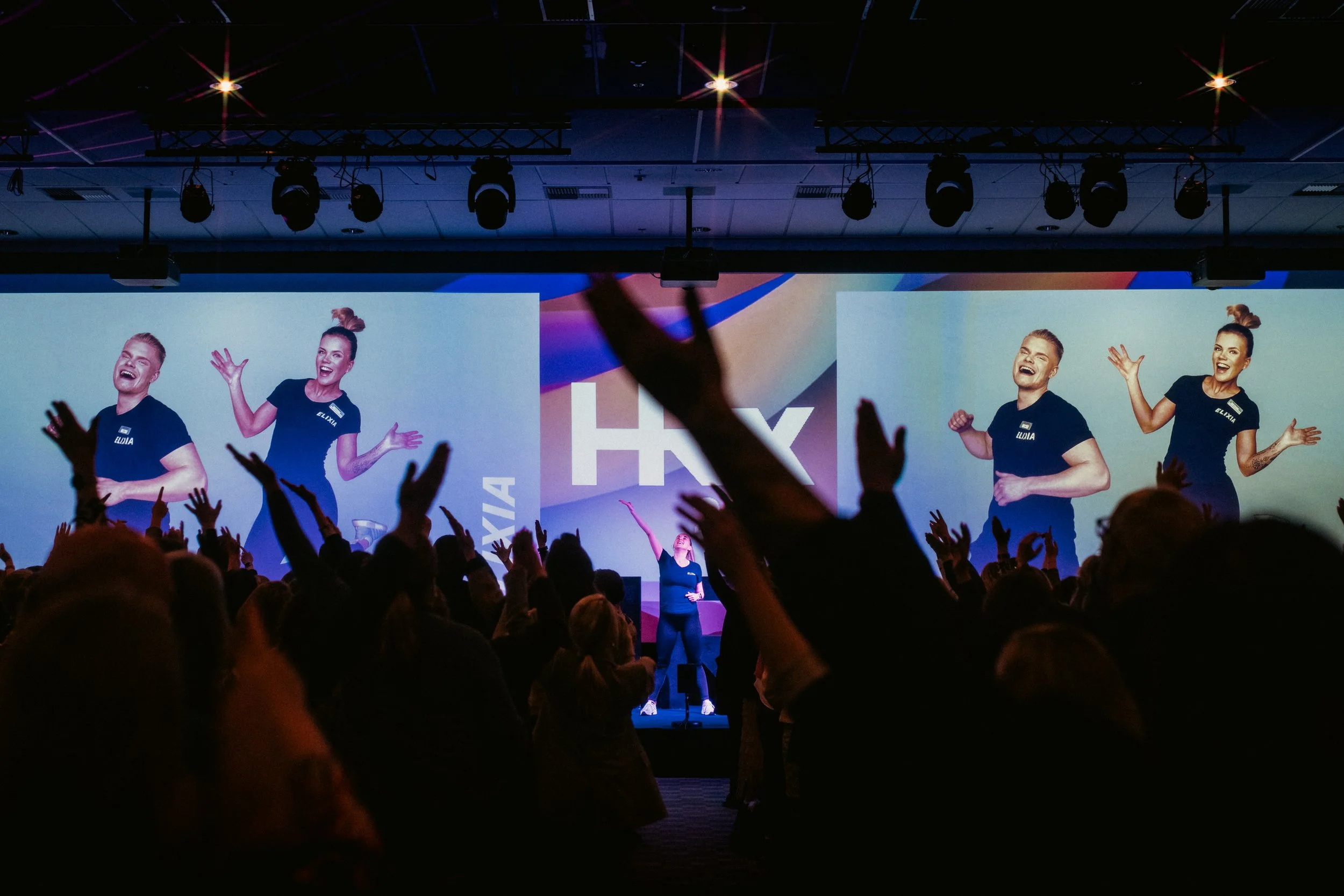 Tapahtuma, jossa ihmiset juhlivat ja nostavat käsiä ylös salissa olevalla lavalla, jossa on suuri näyttö taustalla ja kaksi videota.