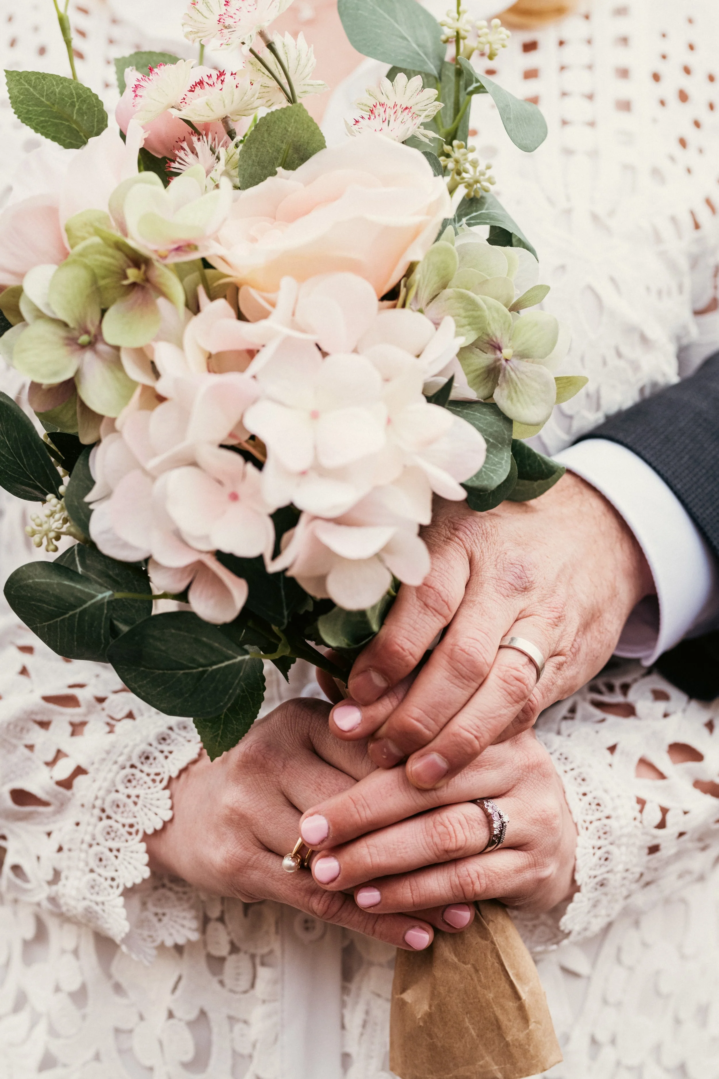 Kaksi ihmisen kättä, joissa on vihkisormukset, pitäen kukkaterästä, jossa on vaaleanpunaisia ja valkoisia kukkia, kuten hortensioita ja pionin kaltaisia kukkia.