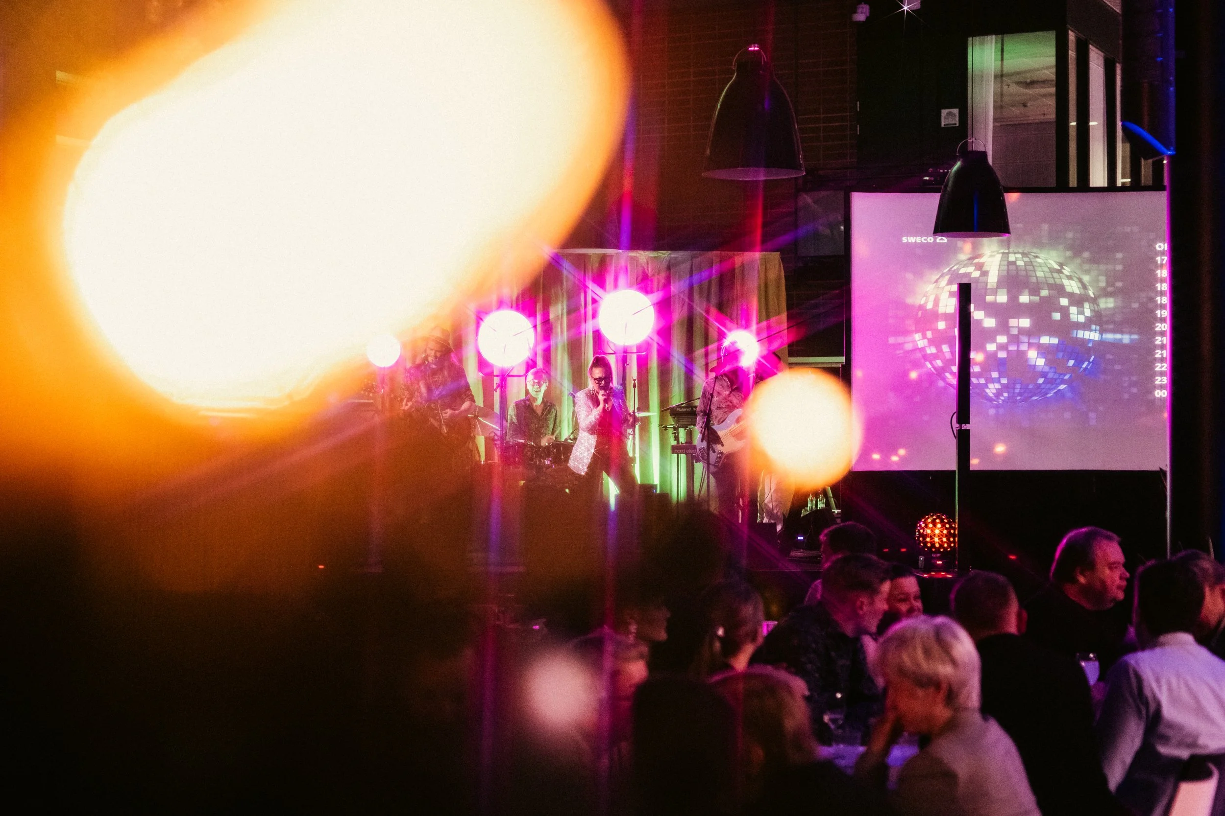 Musiikki esiintymässä baarissa tai ravintolassa, valoefektit ja suuret kellot taustalla, ihmisiä istumassa pöydissä seurustelemassa.