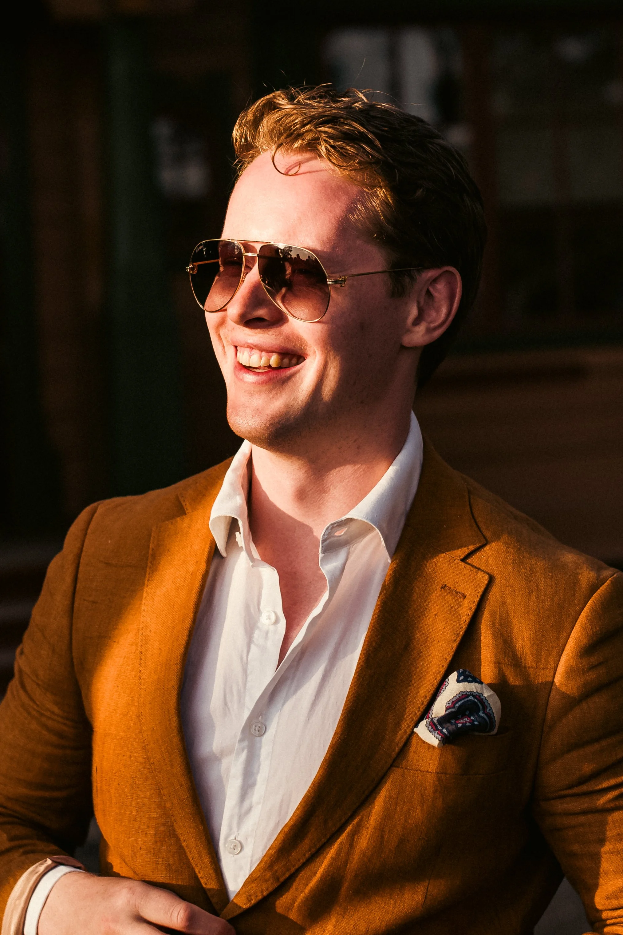 Henkilö hymyilee, hänellä on tummat auringonvarjostuslasit, ruskea sedaanimallinen puku ja valkoinen kauluspaita. Taustalla on hämärää ja lämpimän sävyistä valoa.