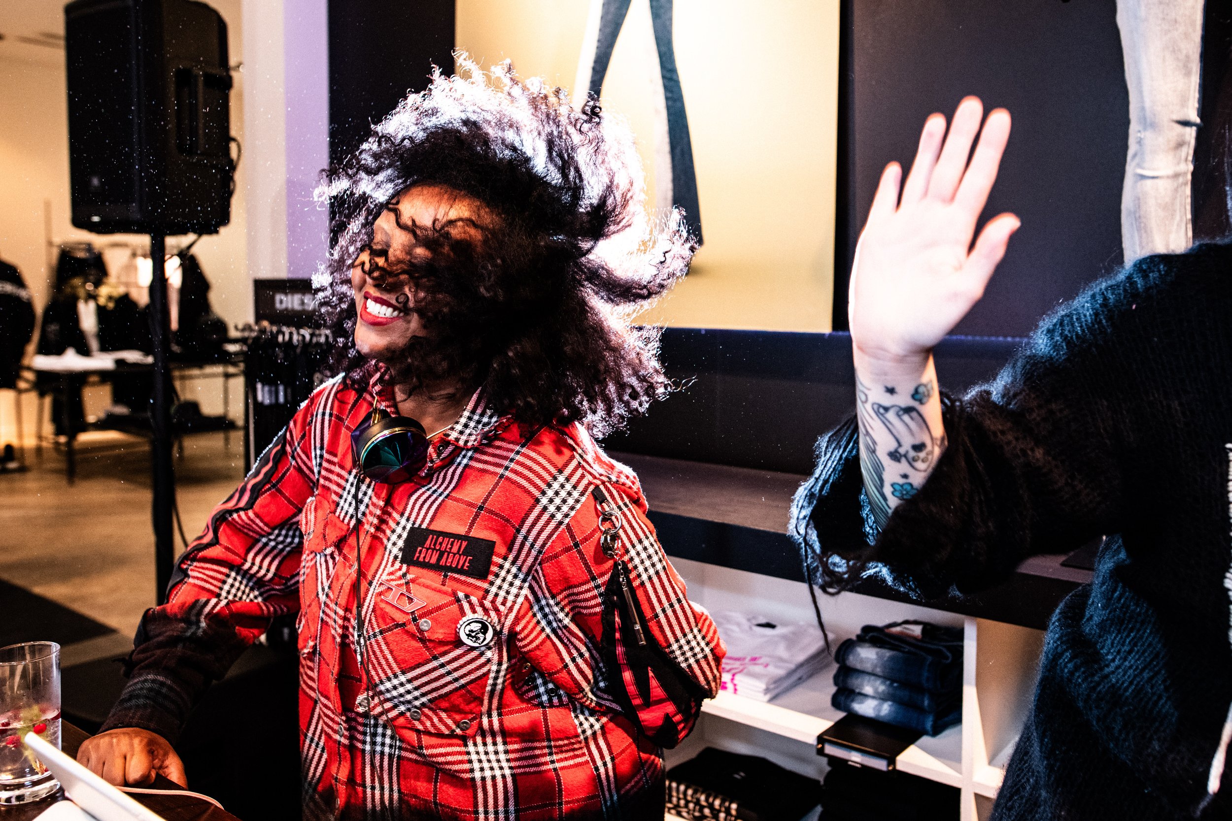 Nainen hymyilee ja tanssii, hänen hiuksensa ovat liikkuvat ja hänellä on kuulokkeet kaulassaan. Hän on pukeutunut punaisiin ja mustiin ruutupaitoihin, päällä on nimilappu ja koristeita. Hän on sisätiloissa, taustalla on tehtävänampi ja muita ihmisiä.