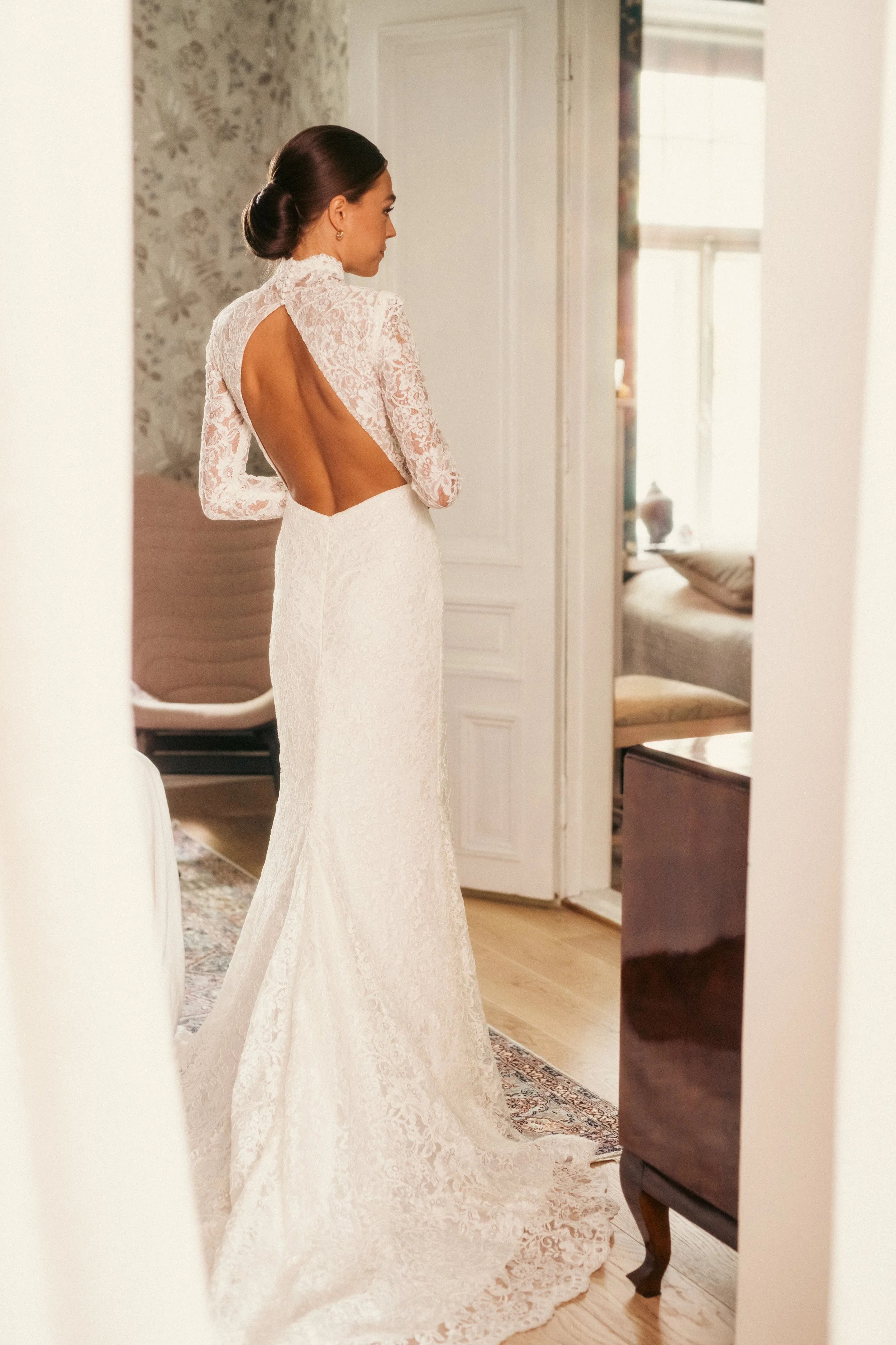 Nainen mekossa kuvassa sisältä, paljastaa selkänsä, on kiharatukkainen ja hiukset nutturalla, kuvataan sisätiloissa vanhan tyylisessä huoneessa.