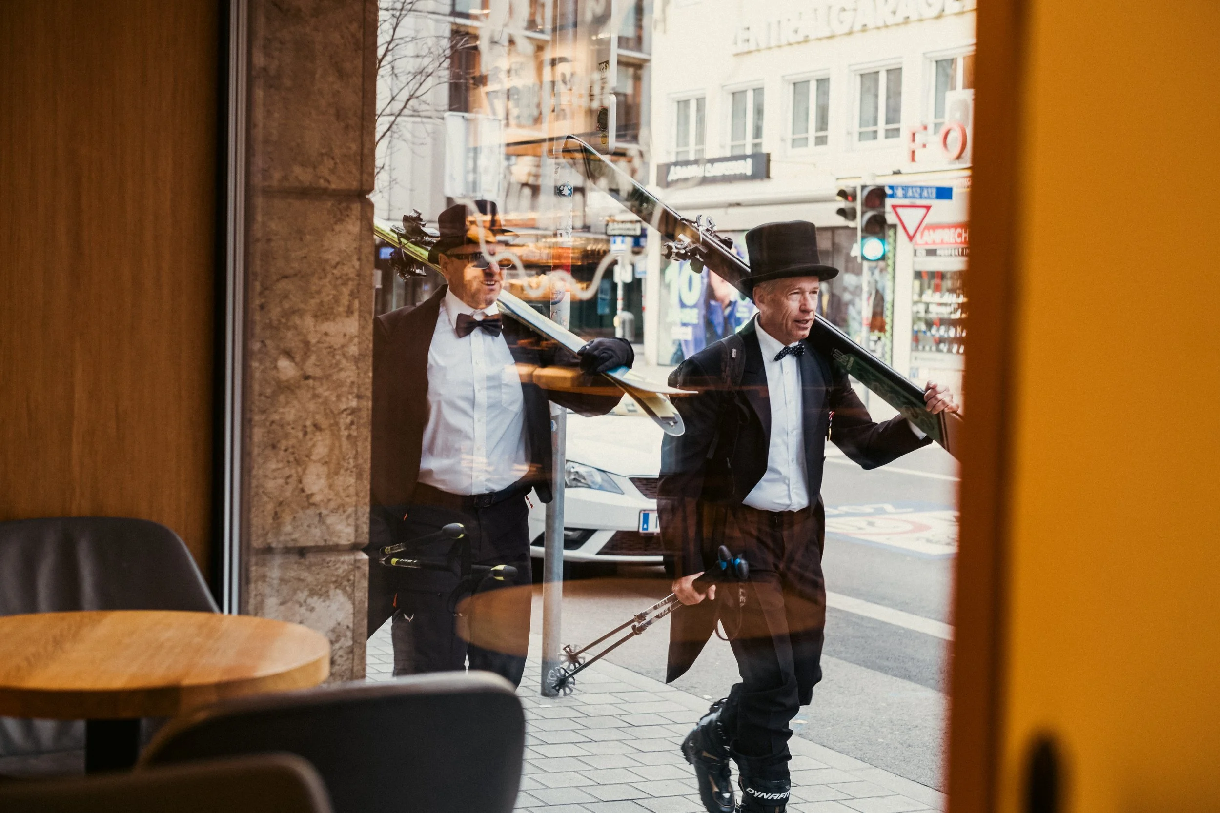 Kuva, jossa kaksi miestä kävelee kadulla, pukee frakit ja hatut, yksi kantaa urheilutossia ja sauvoja.