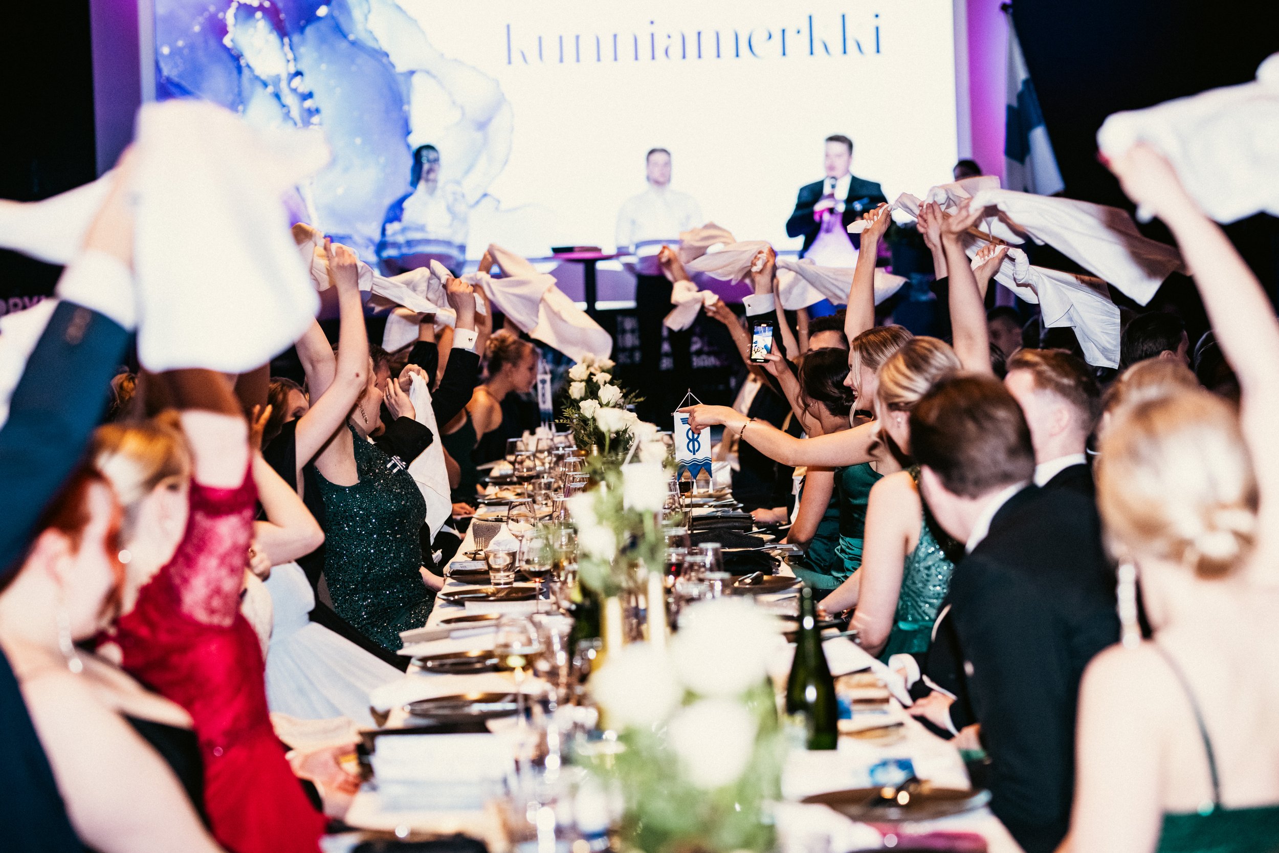 Henkilöitä juhlapöydässä, joissa ihmiset tanssivat ja heiluttavat pyyhkeitä, takana suuri näyttöruutu ja kutsuvieraita.