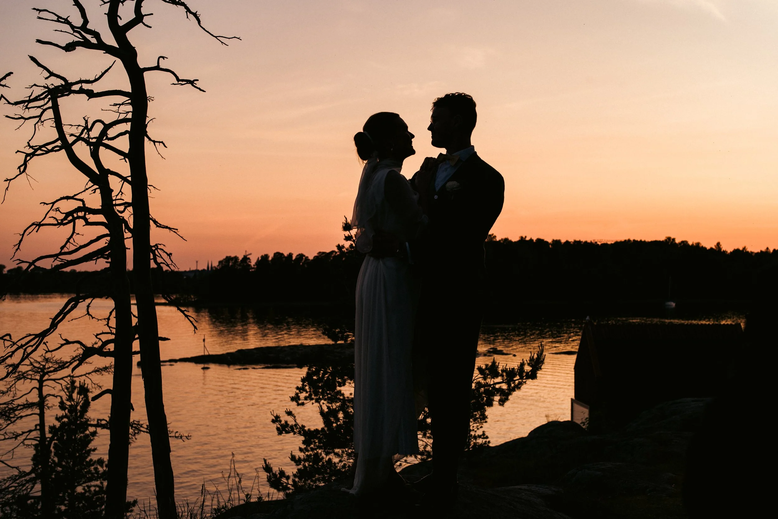 Pariskunta ihailee Alps-järven taakse laskevaa aurinkoa, taustalla oliivipuita, tyhjä talo ja laiva vedessä, täysin varjostettu liitua ja ihmisiä sisältämättömän siluetin alla.