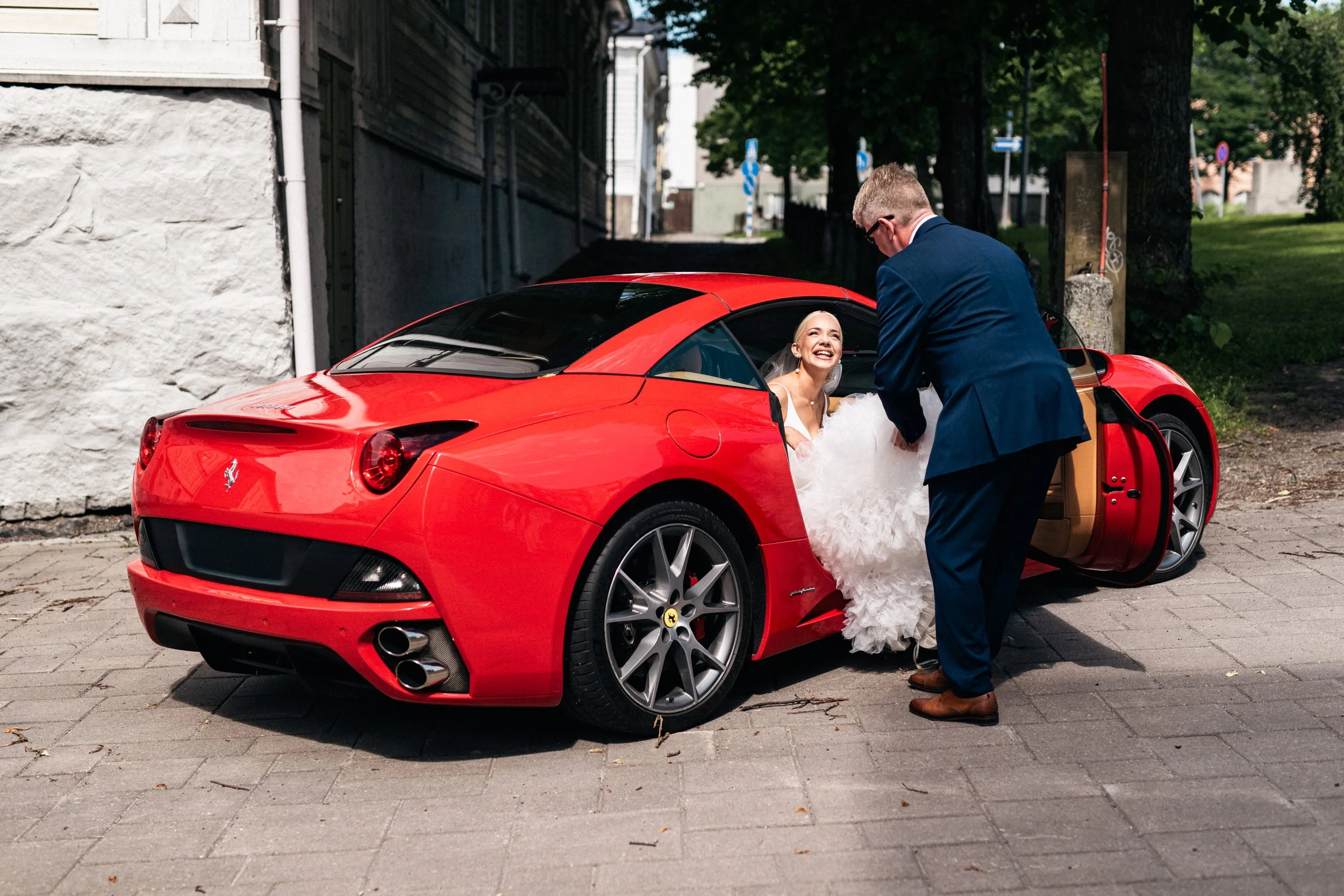 Hääpari poistuessaan punaisesta Ferrari-autosta, jossa morsian istuu autossa hymyillen, morsian pukeutuneena valkoiseen häämekkoon, ja sulhasen pukema tummansininen puku, kadulla päivänvalossa.