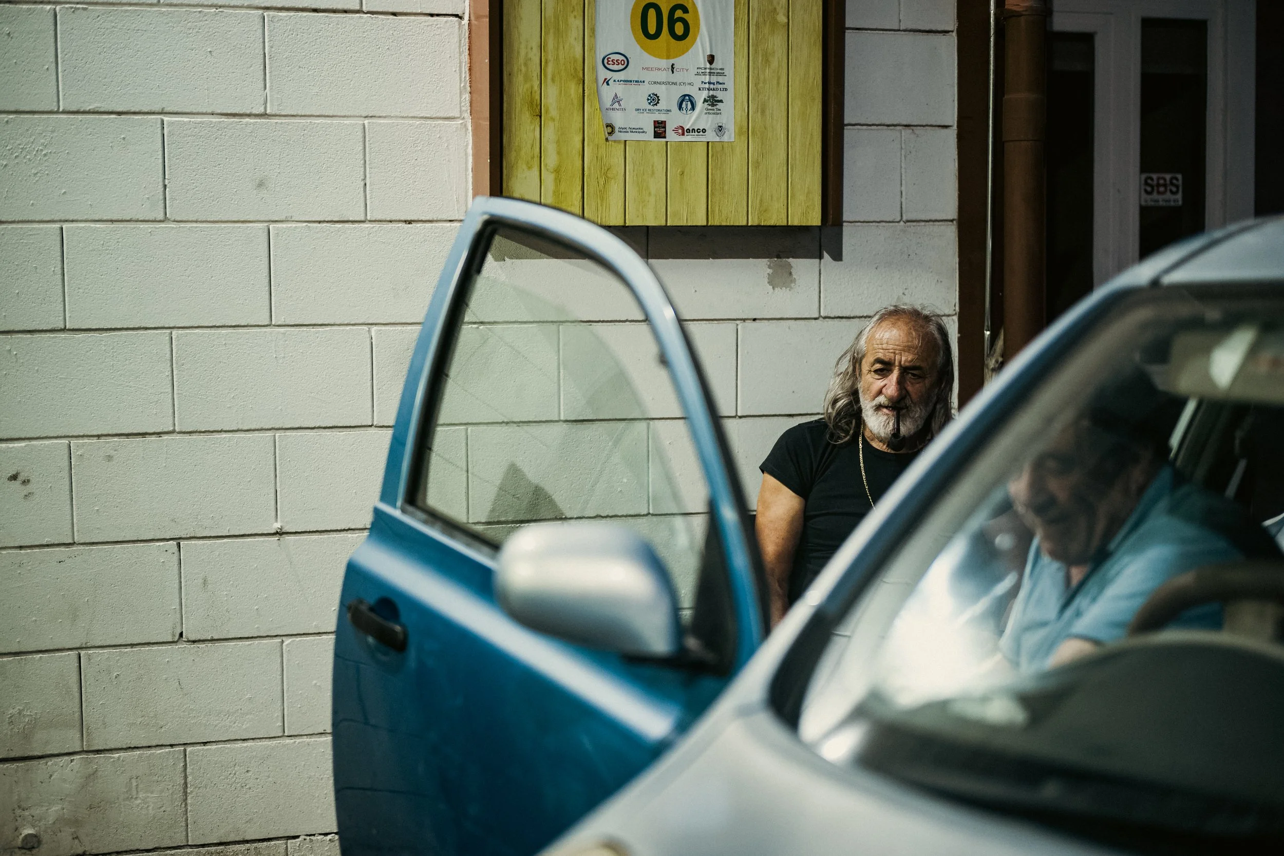 Kaksi miestä, toinen istuu autossa ja toinen seisoo vieressä, viereisessä pysäköintialueella, vanhan tiilitalon edessä