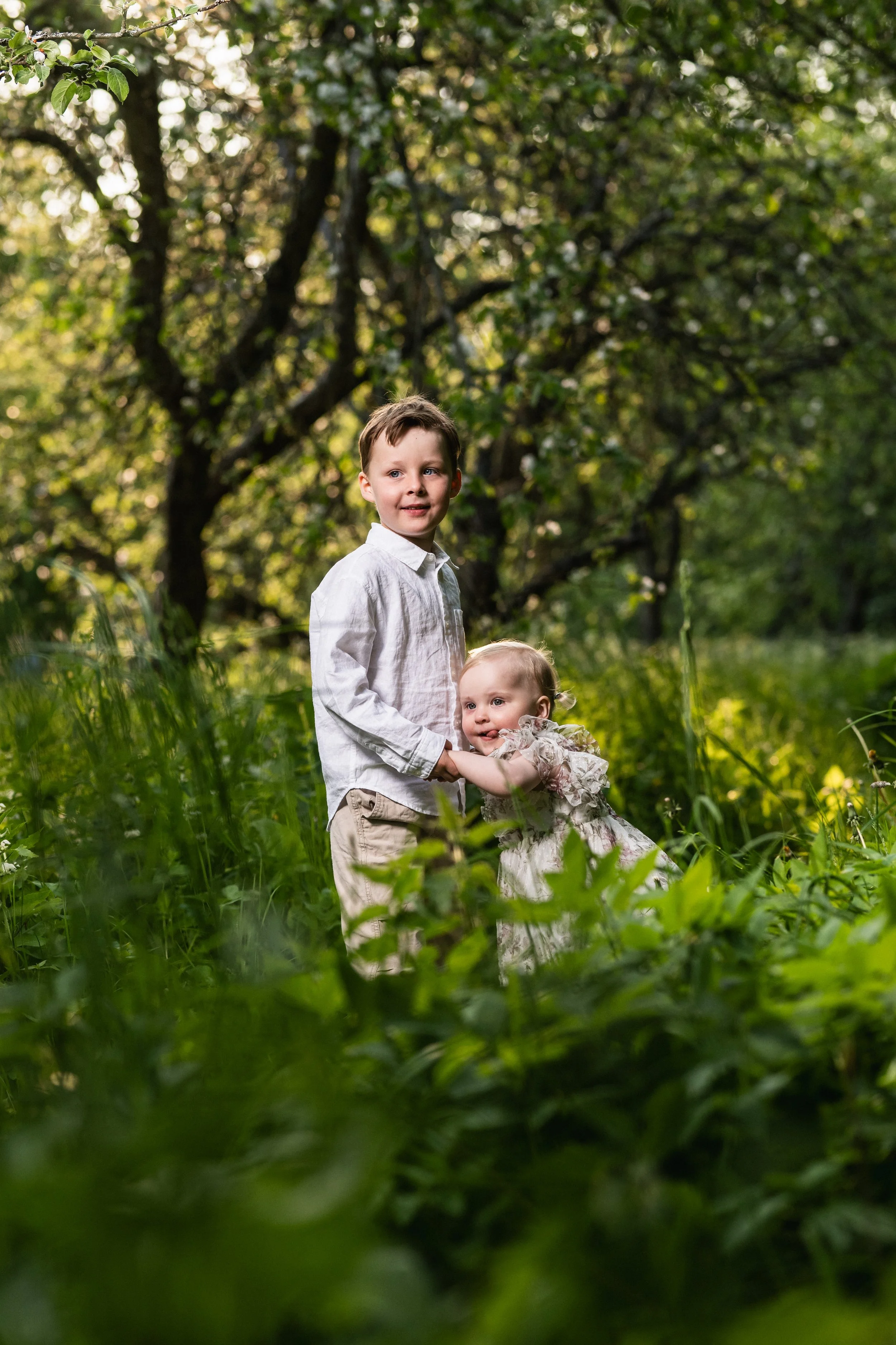 Kaksi lasta, poika ja tyttö, kävelee metsässä kasvatuskasvillisuuden keskellä, taustalla puita ja vihreitä lehtiä.