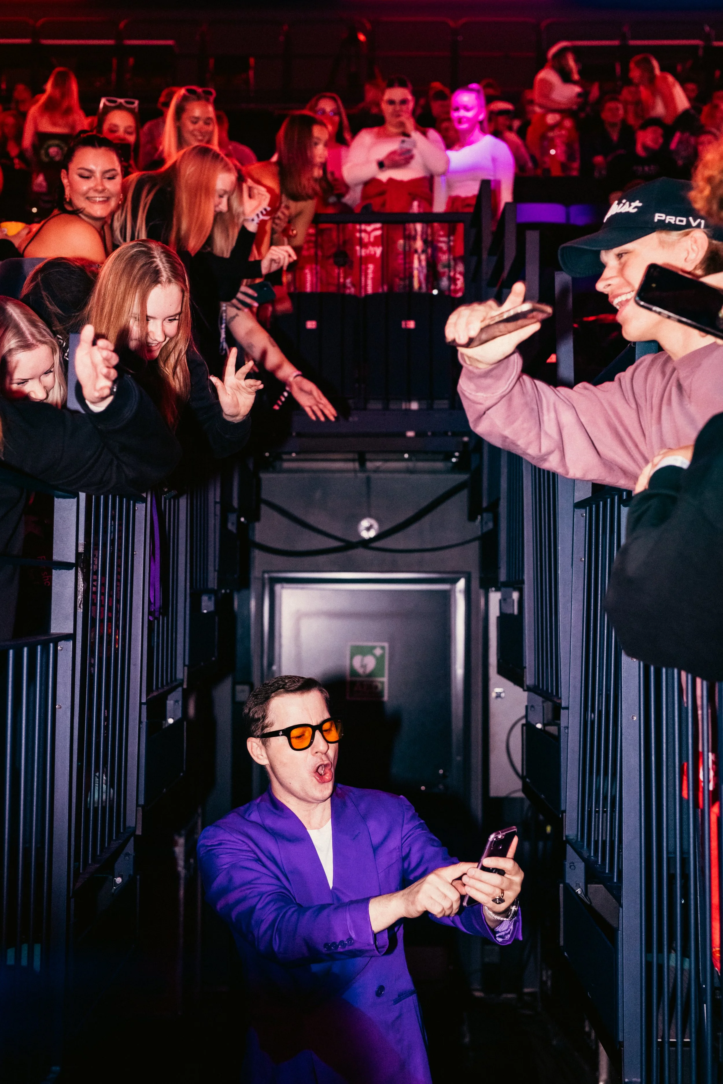 Nuori mies, jossa tummat hiukset, puvuntakki ja keltaiset lasit, seisoo ja katsoo puhelintaan, joka hänellä on kädessään, ja hänen ympärillään on innostuneita nuoria ihmisiä kasinoalueella.