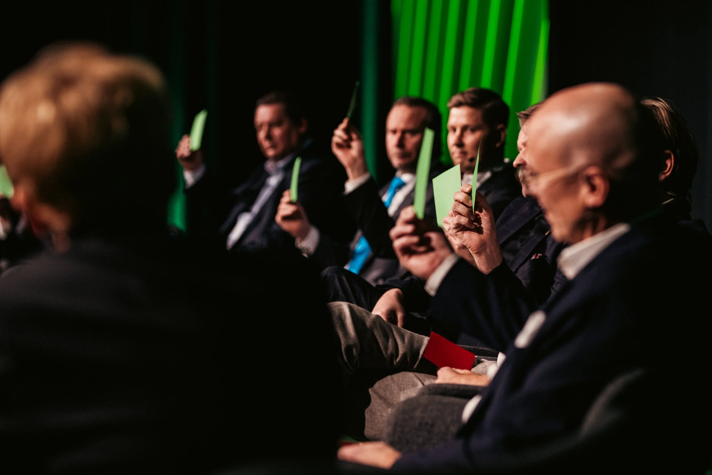 Ihmisiä istumassa, osa pitää vihreitä ja punaista merkkiä, ilmeiltään vakavia, taustalla vihreitä valoja.