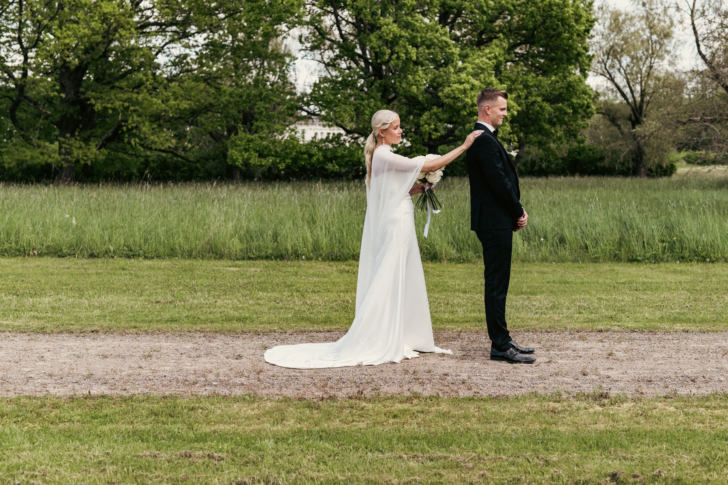 Hääpari seisoo ulkona nurmikentällä, kulta ja valkoinen hääpuku, juhlava puku, vihreitä puita taustalla.