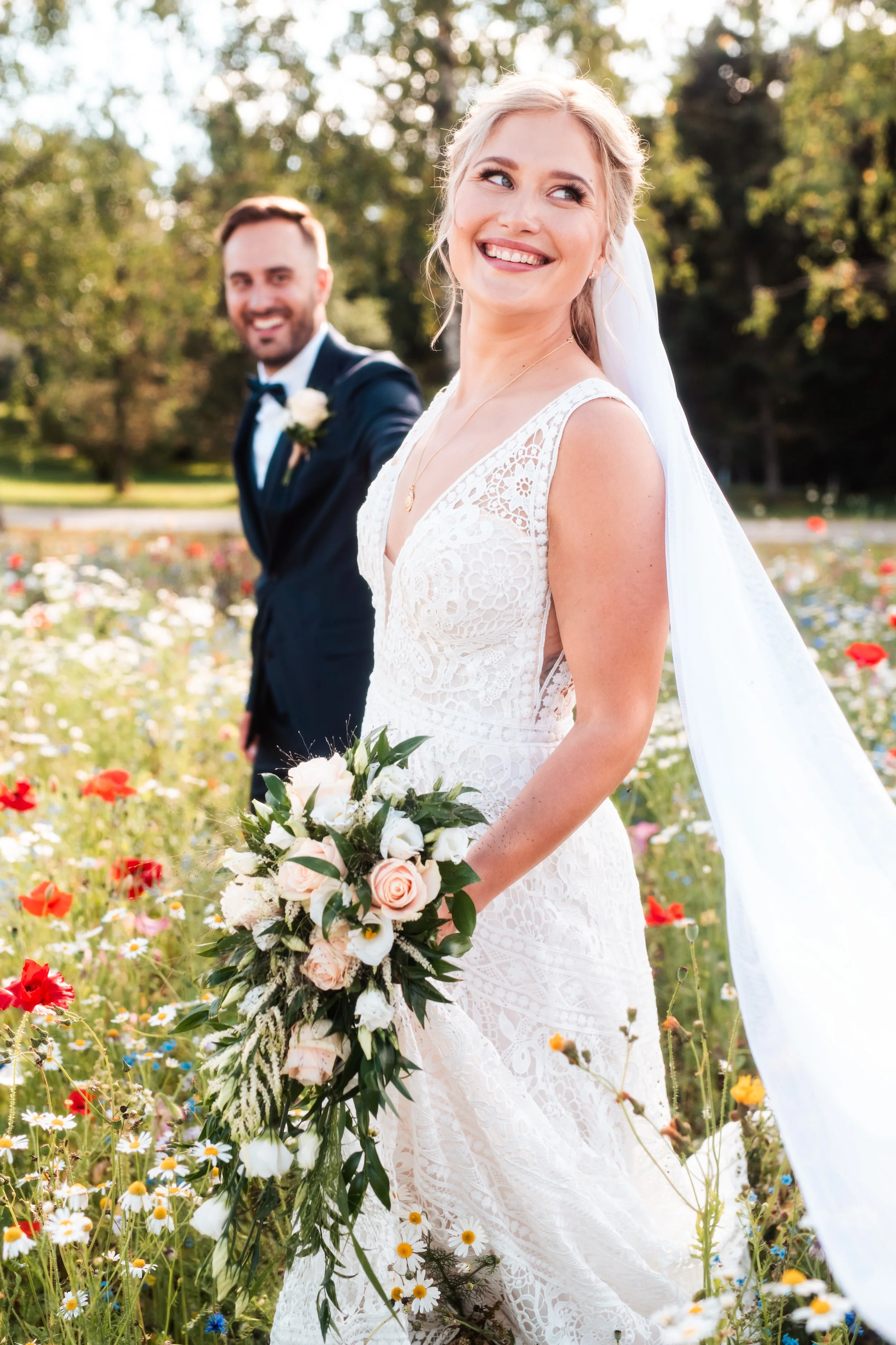 Vihkisormuksen ja hääpuvun kanssa oleva morsian hymyilee ja poseeraa kukkakedolla, ja häntä seuraava sulhanen hymyilee taustalla.