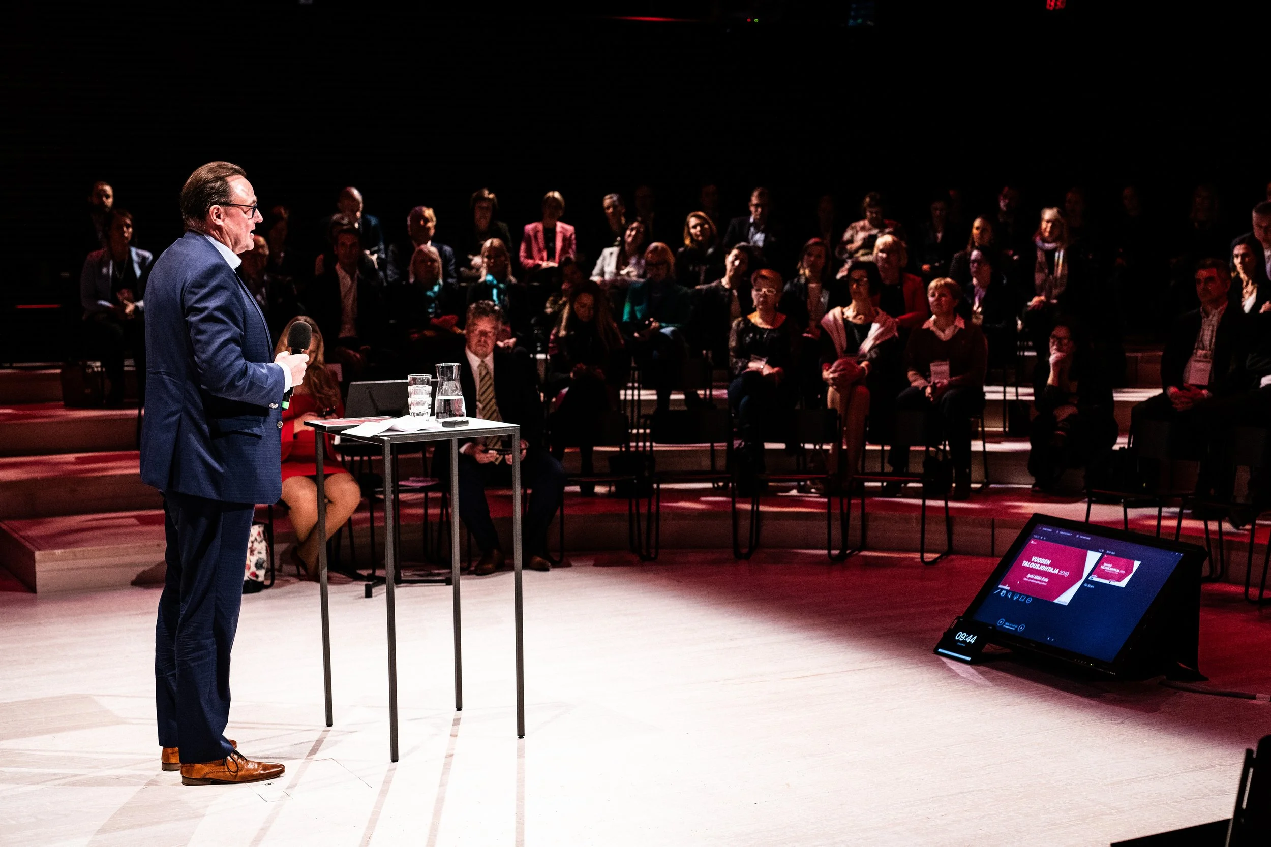 Mies puhuu mikrofonilla lavalla, yleisö istuu katsomossa ja katsoo häntä, vilkkuva sähköinen näyttö säädetyissä valoissa.