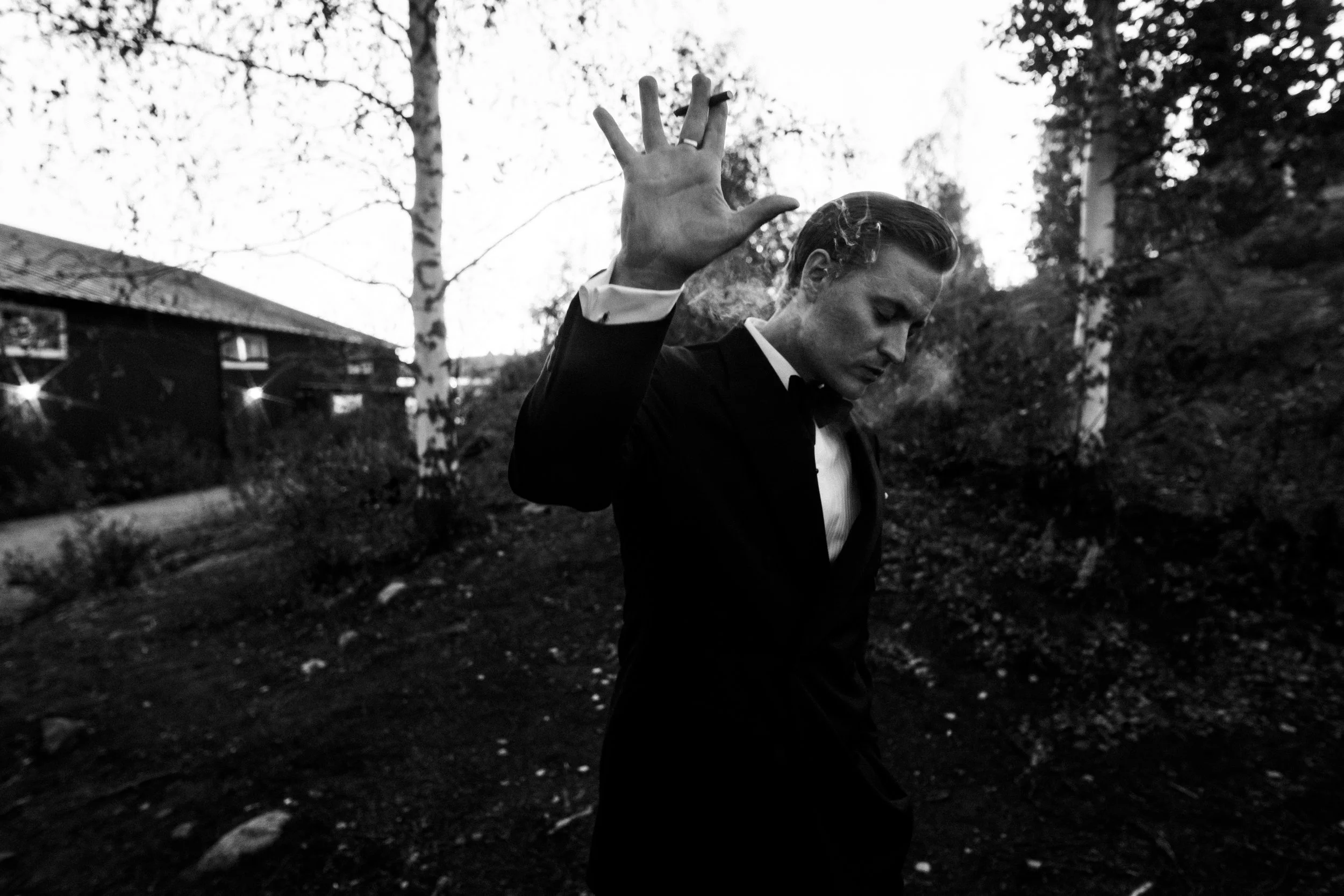 Mies pukeutuneena smokkiin, kädellään tehdessään jännittynyttä liikettä, ulkona pimeässä ympäristössä puita ja rakennuksia.