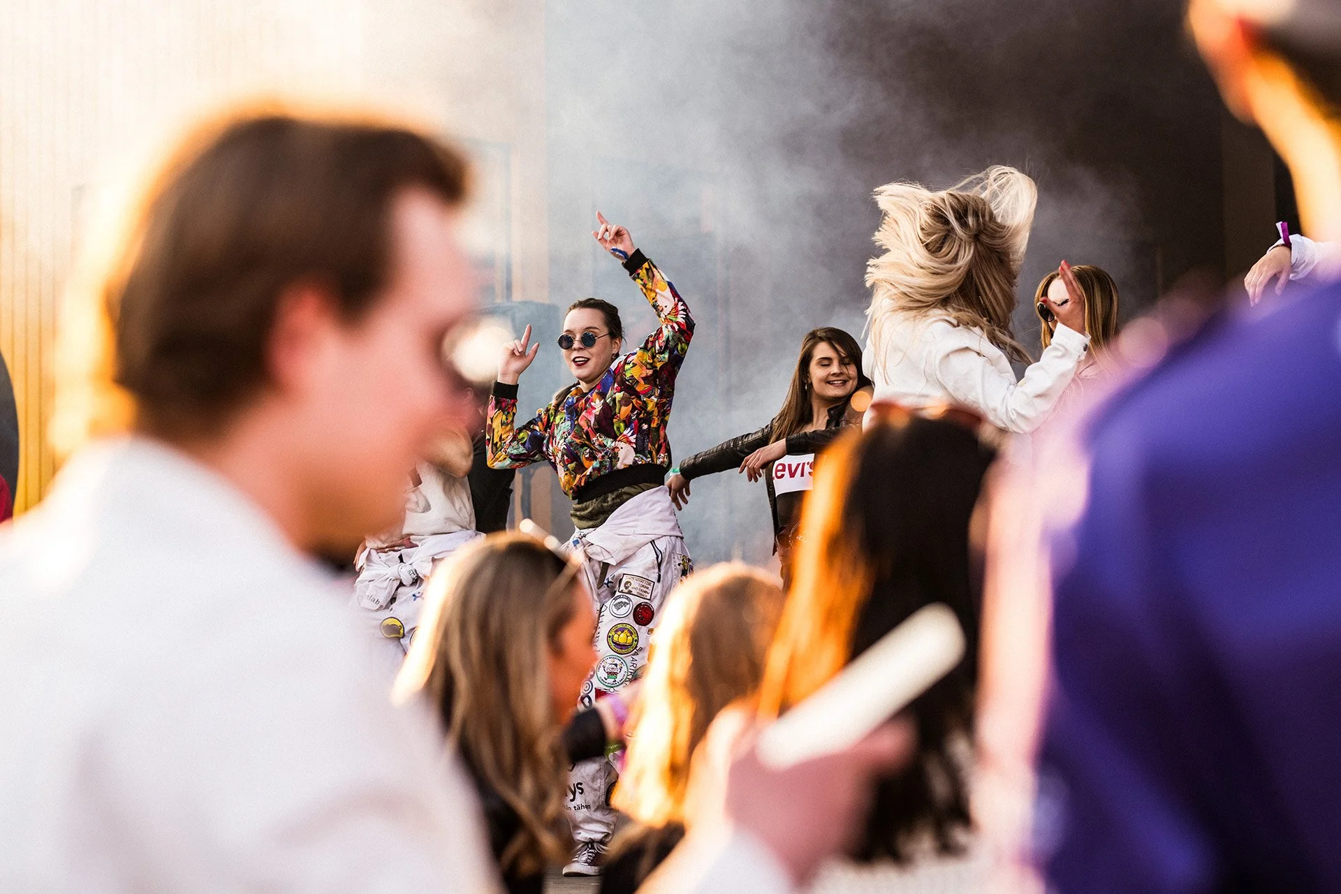 Nuori ryhmä ihmisiä tanssimassa ja juhlimassa, iloisia kasvoja, värikkäitä vaatteita, esiintymis- tai juhlatilassa.