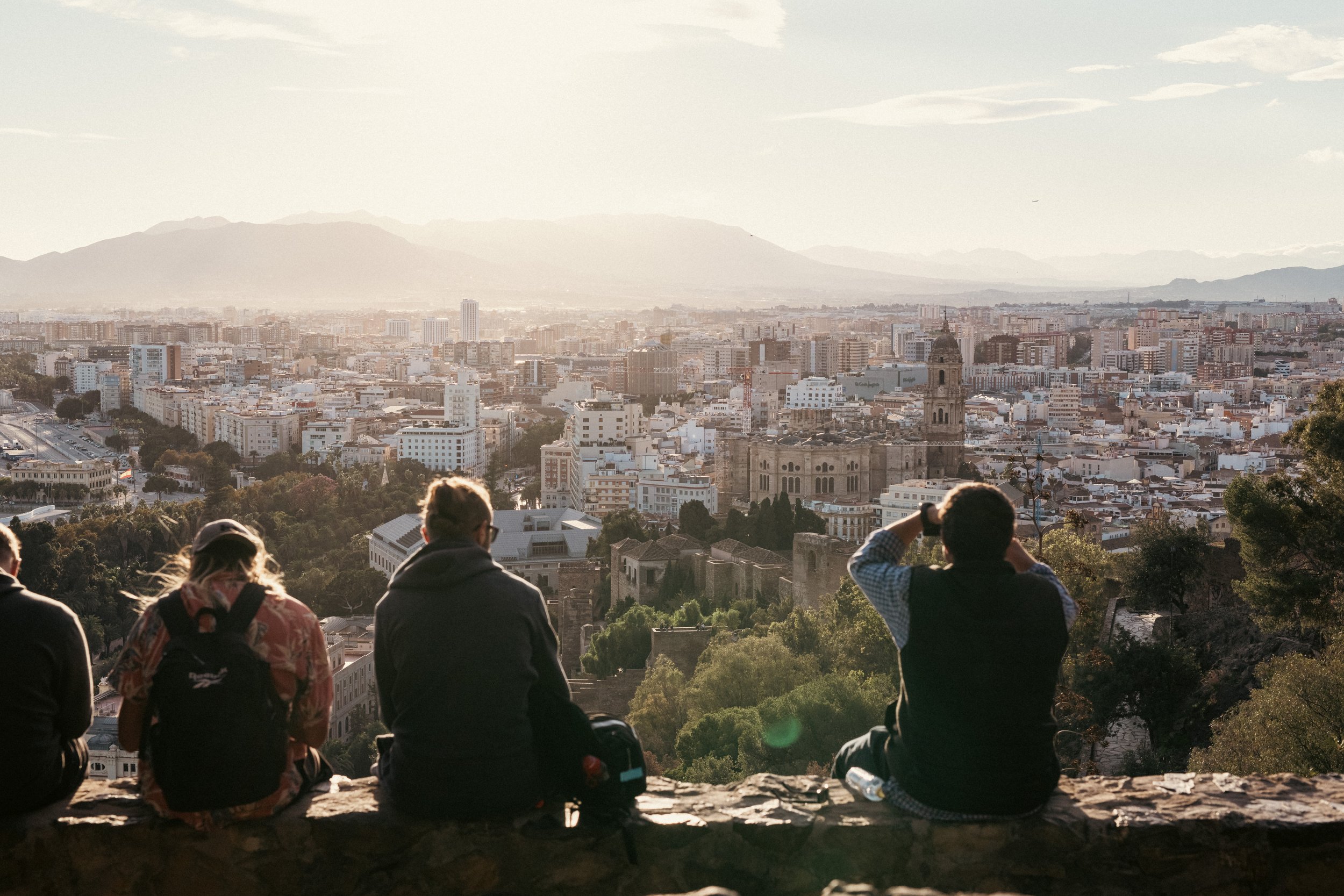 Neljä ihmistä istuu kiviseinällä katsellen kaupunkimaisemaa auringonlaskun aikaan, taustalla kaupunkia ja vuoria.