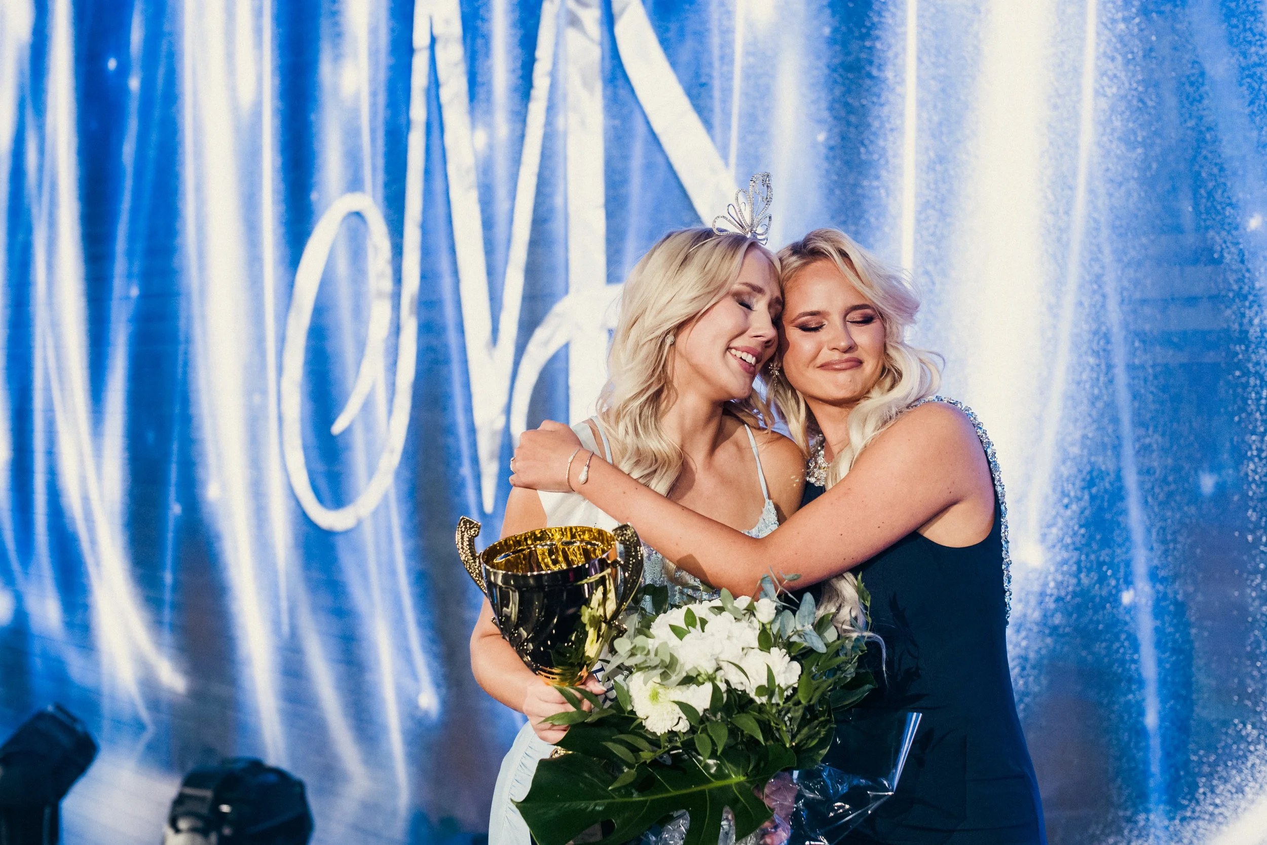 Kaksi naista halaavat ja iloitsevat voitosta, toinen pitää kukkakimppua ja pokaalia, heillä on iloiset ilmeet ja taustalla sinertävä verho.