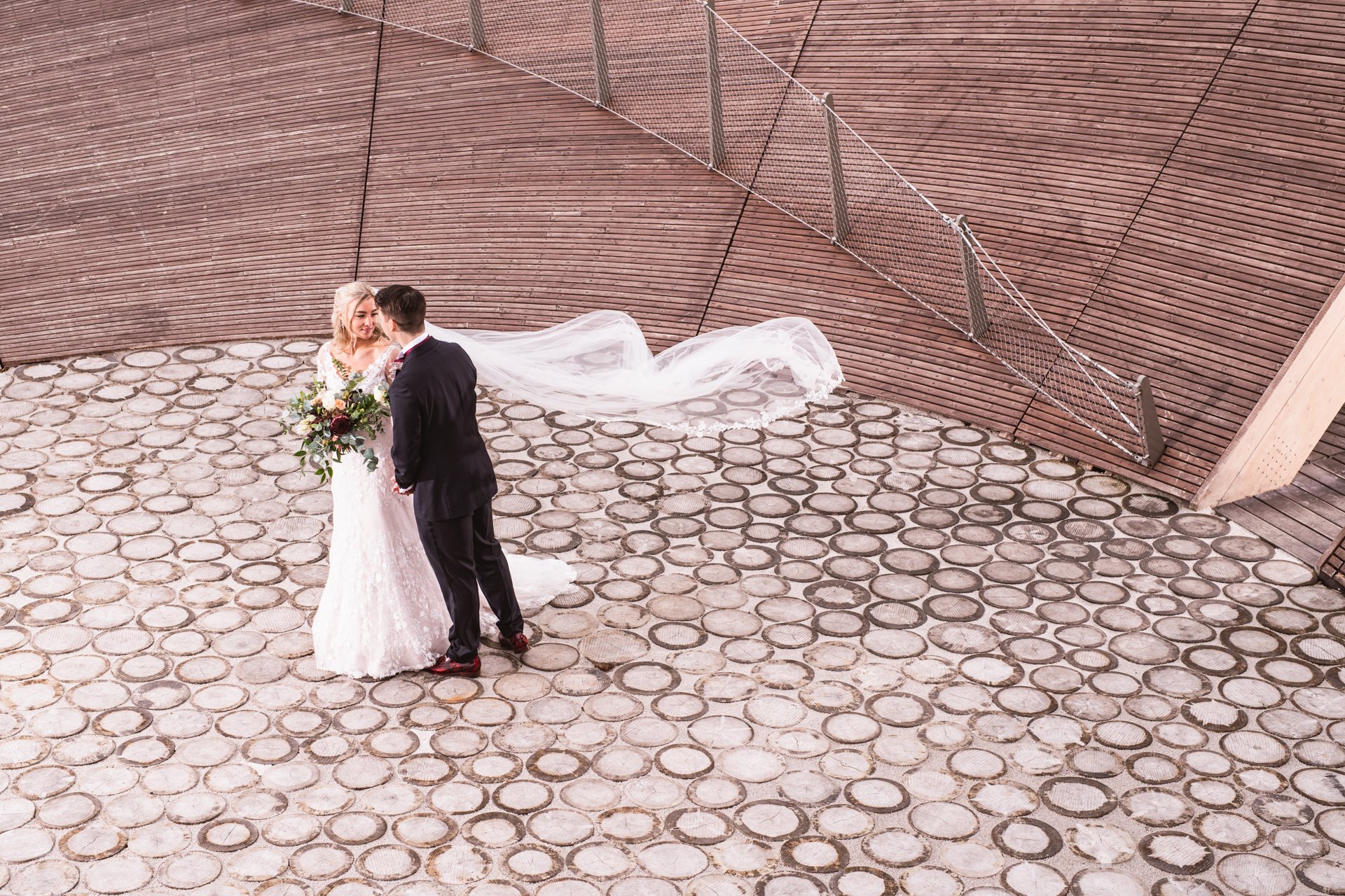 Hääpari tanssimassa ulkona kivetyllä alueella, nainen valkoisessa hääpuvussa ja mieheni tummassa puvussa, nainen pitää kukkakimppua.