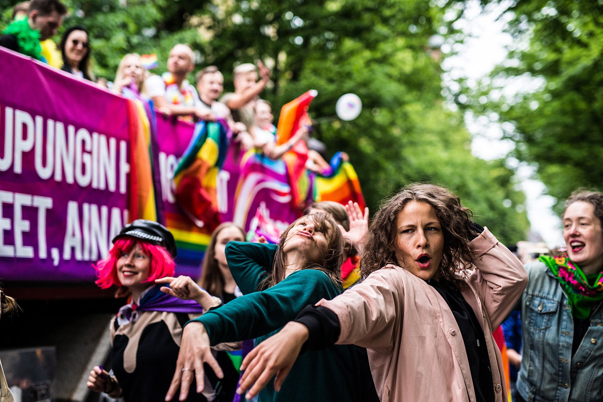 Ihmiset osallistuvat pride-kulkueeseen, joilla on värikkäät sateenkaariliput ja aseet. Taustalla näkyy suuri sateenkaarilippu ja vihreitä puita.