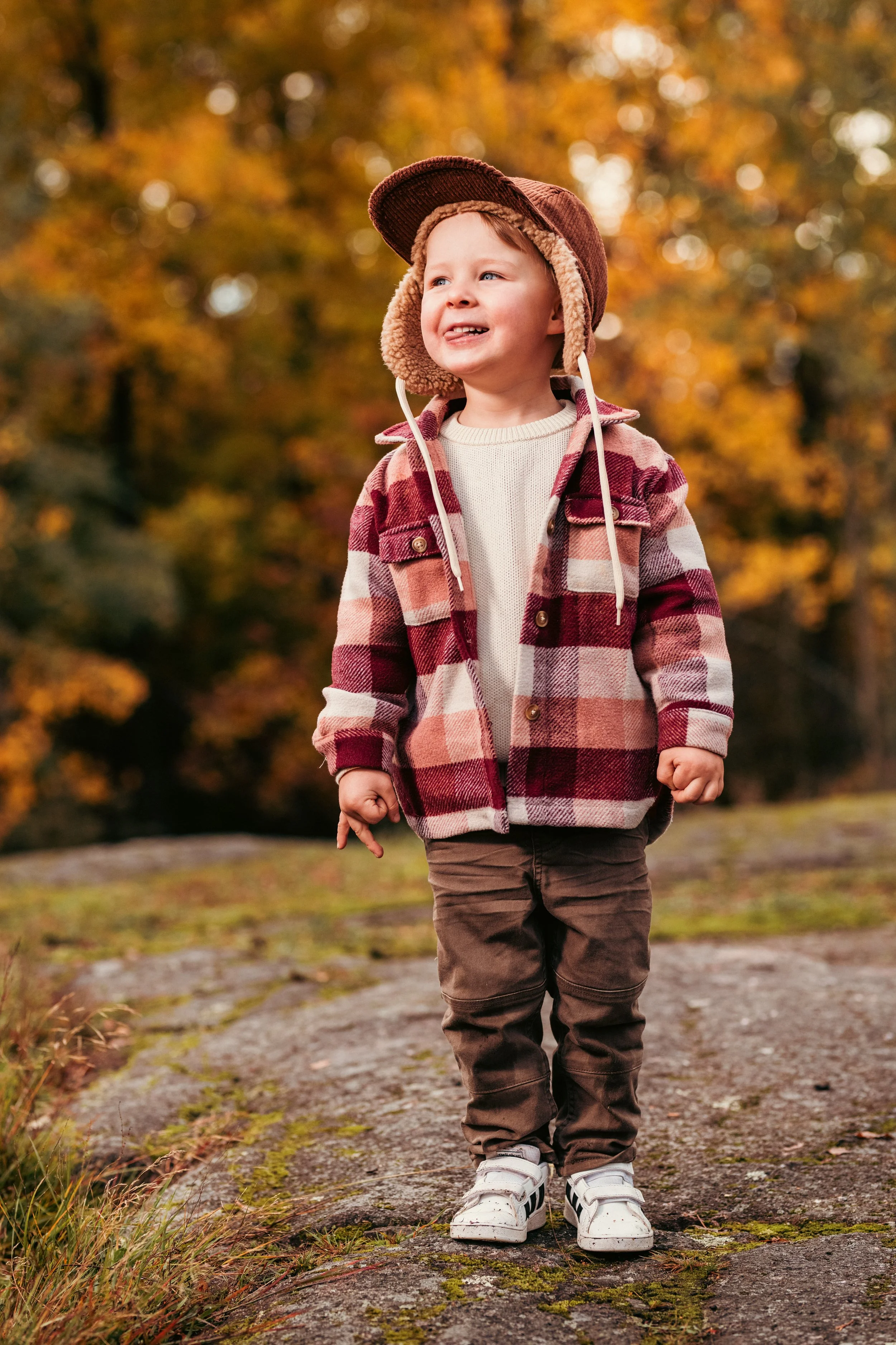 Pieni poika ulkona syksyllä, ruskeassa ja punaisessa ruutupaitatakin, takin huppu ja ruskea lierihattu päässä, hymyilee ja katsoo pois kamerasta, syksyinen metsä taustalla.