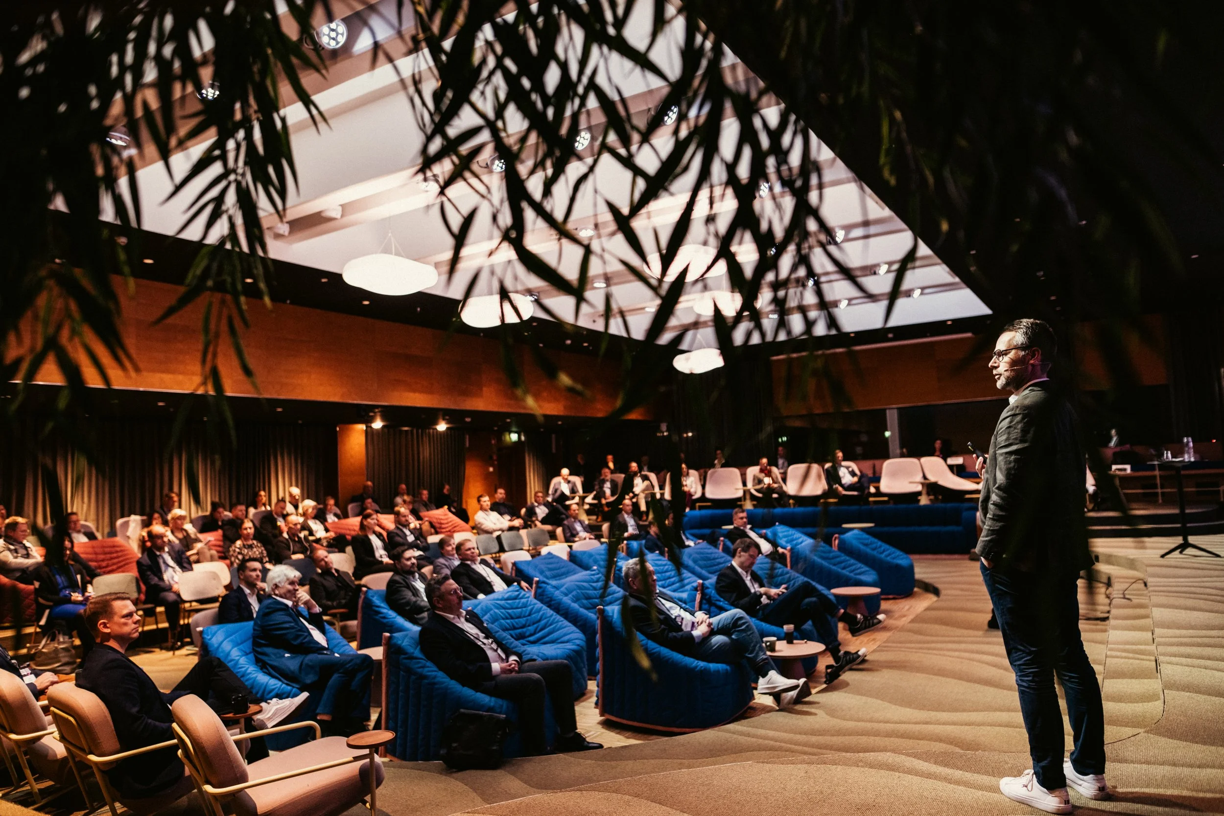 Konferenssali pressenttiä kuuntelemassa ihmisiä, katsovat eteenpäin, osa käyttää pöytäkoneita. Yleinen tunnelma on ammatillinen ja keskittynyt.
