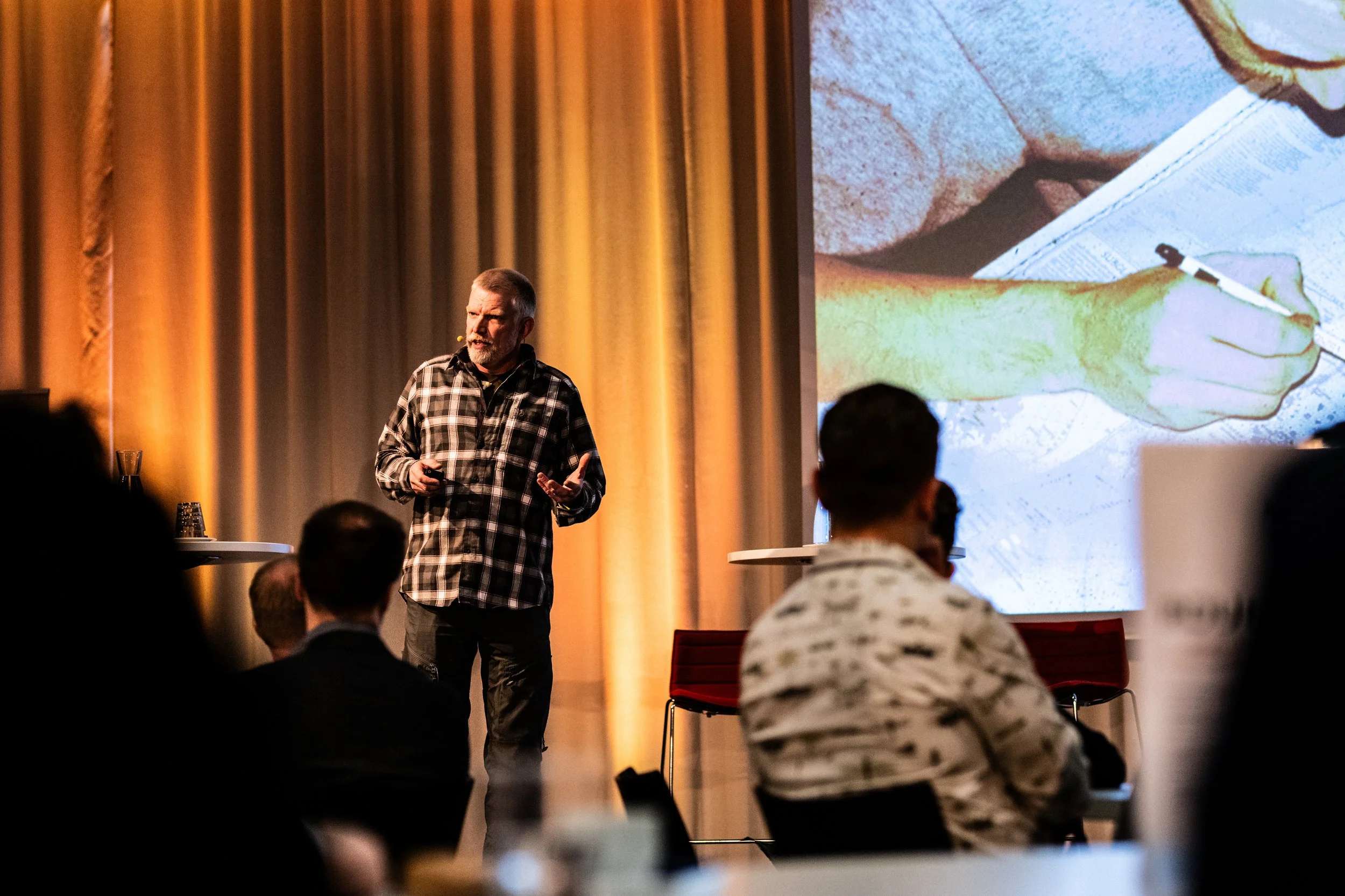 Mies puhuu lavalla, taustalla suuri projektiokuva ja haaleat keltaiset verhot, paikalla istuu ihmisiä.»
