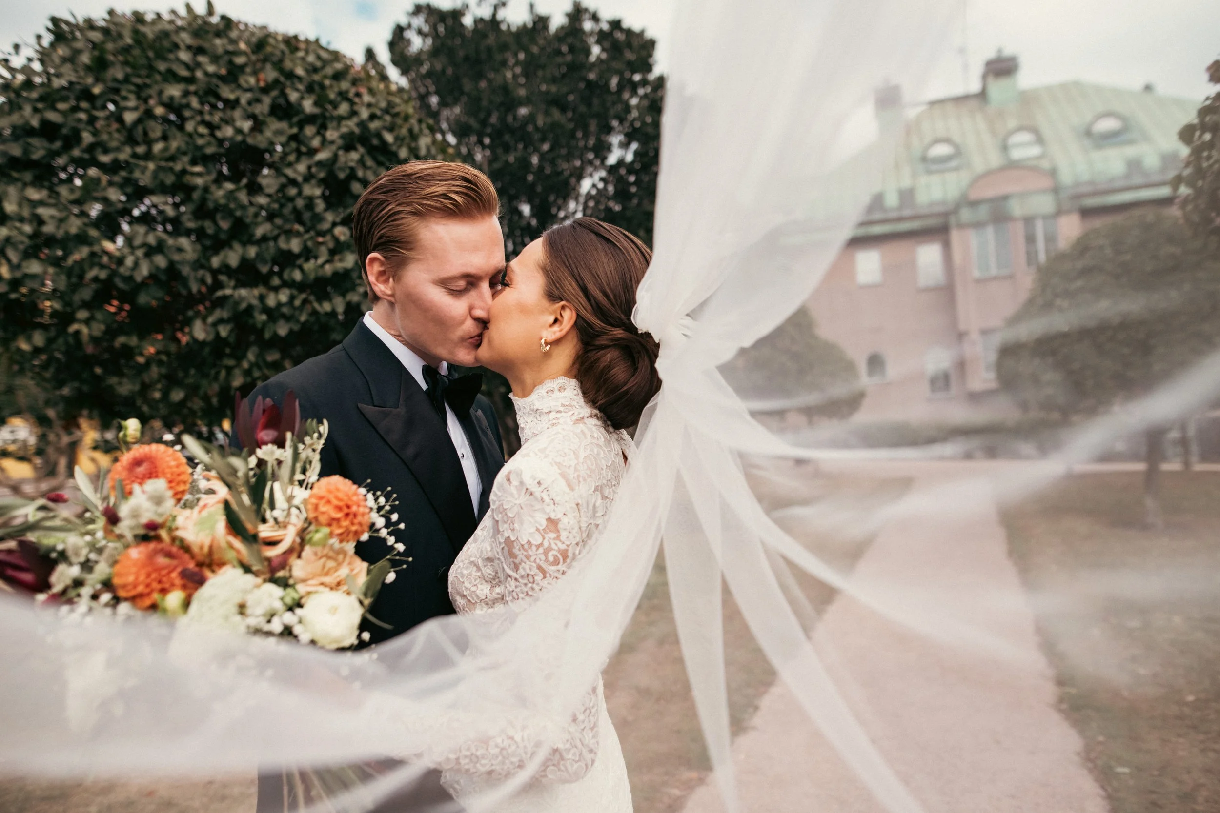 Hääpari suutelee toisiaan ulkona, nainen pitelee kukkakimppua, taustalla puita ja rakennus.