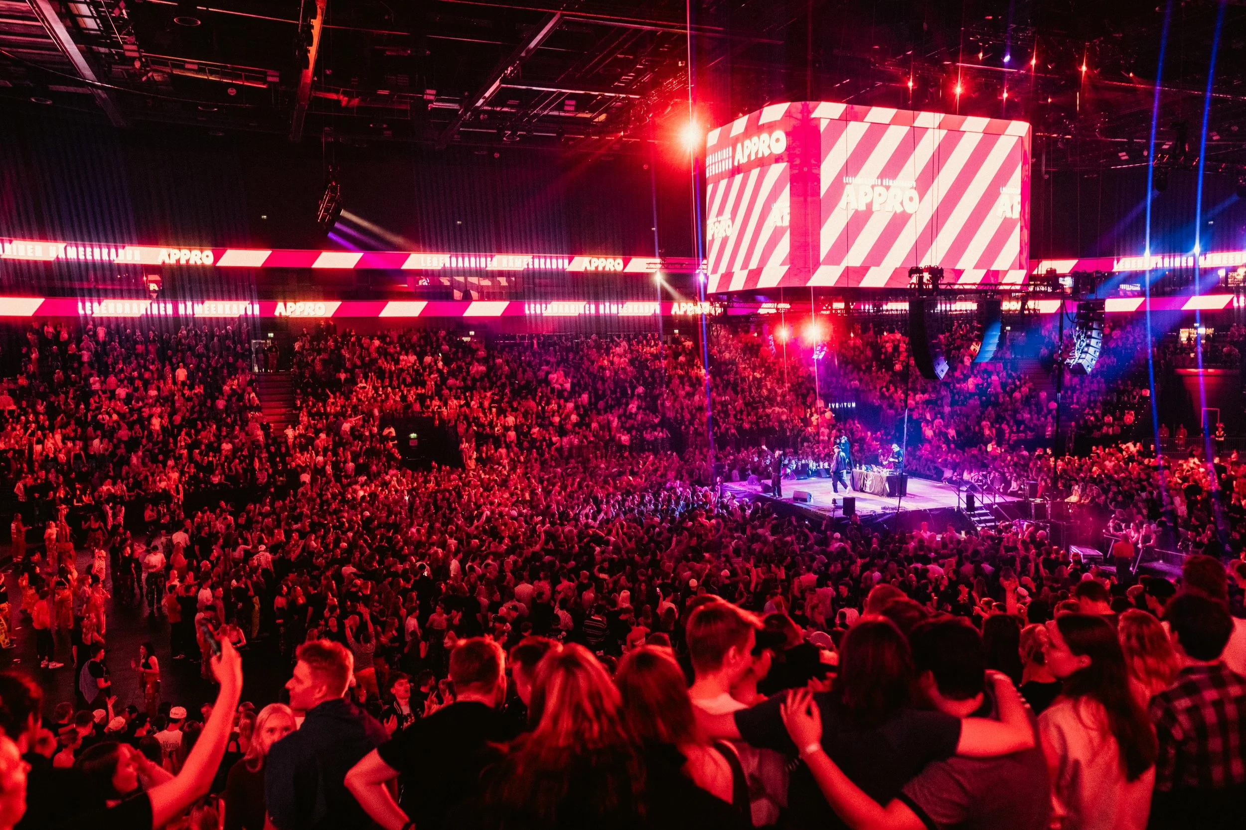 Suuri konserttitapahtuma täynnä ihmisiä, lavalla esiintyy artisti ja taustatykki näytöllä näkyy logo 'APPRO', ympärillä värikkäitä valoja ja valotehosteita.